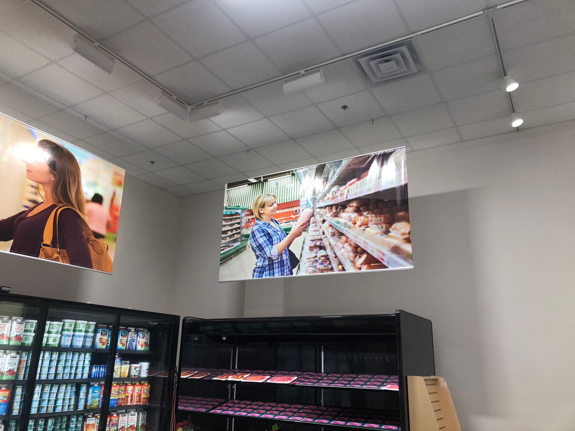 Grocery store interior, displays and advertisements hanging from ceiling. Shelves with products, shoppers featured in ads.