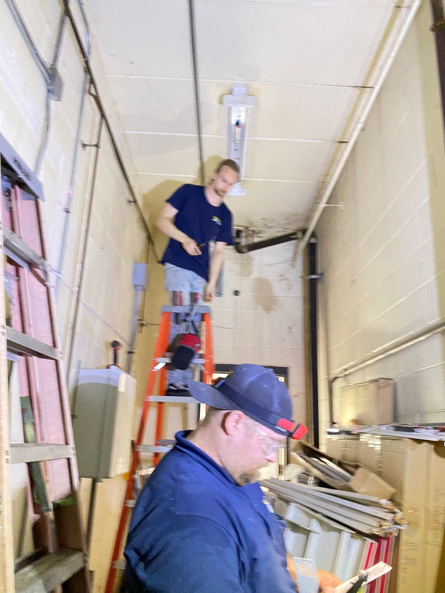 Two men working in a narrow, cluttered room. One on a ladder, the other standing below.