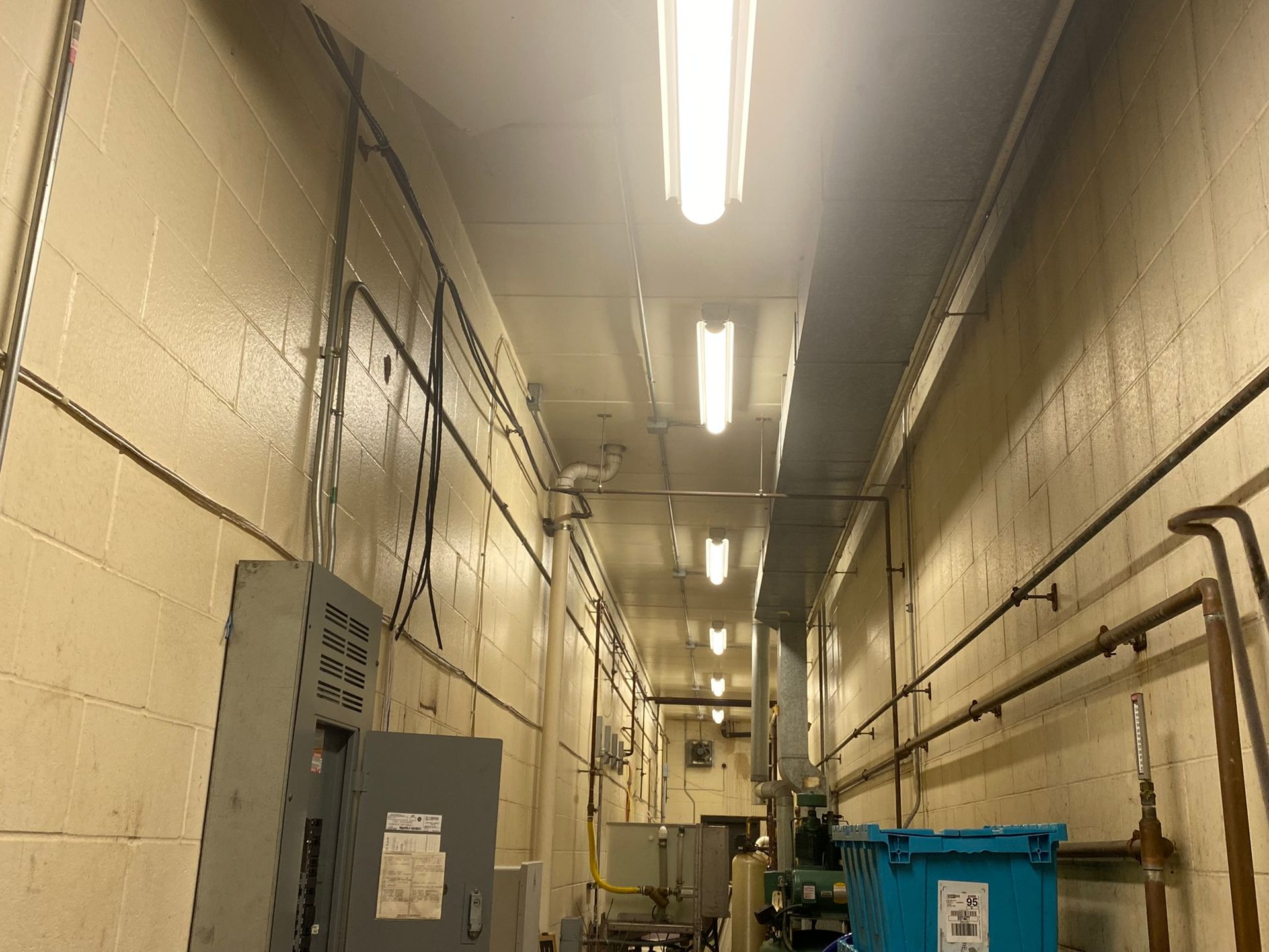 Narrow industrial hallway with electrical boxes, conduits, and overhead lights.