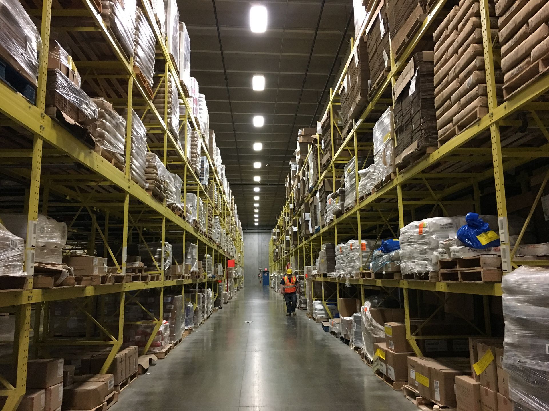 Warehouse interior with tall yellow shelving filled with packaged goods; person in an orange vest walks down the aisle.