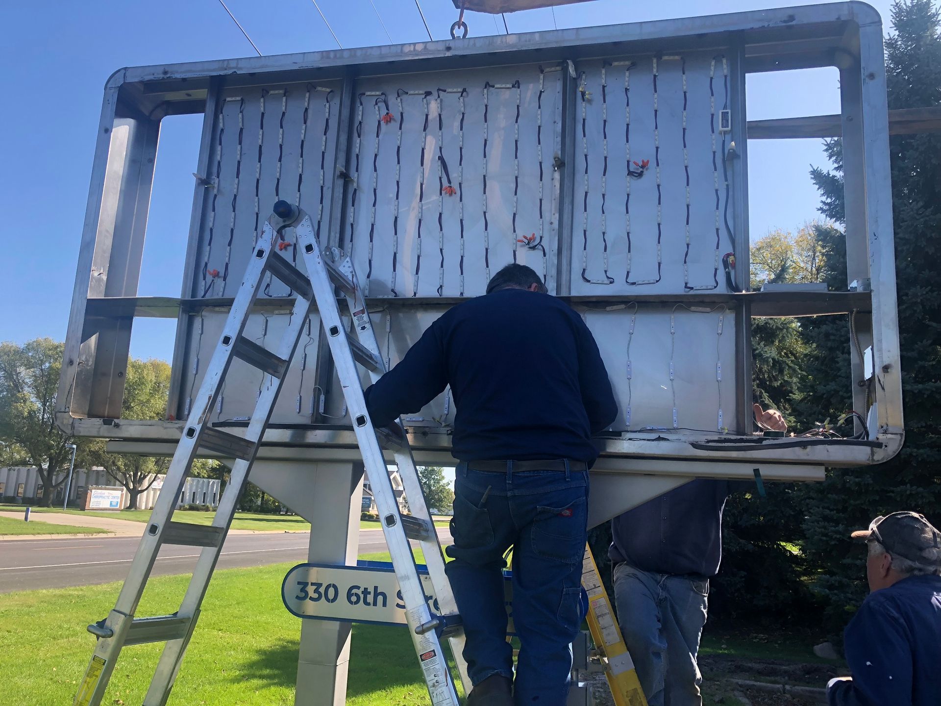 Workers repairing a billboard frame with a ladder, outside on a sunny day.