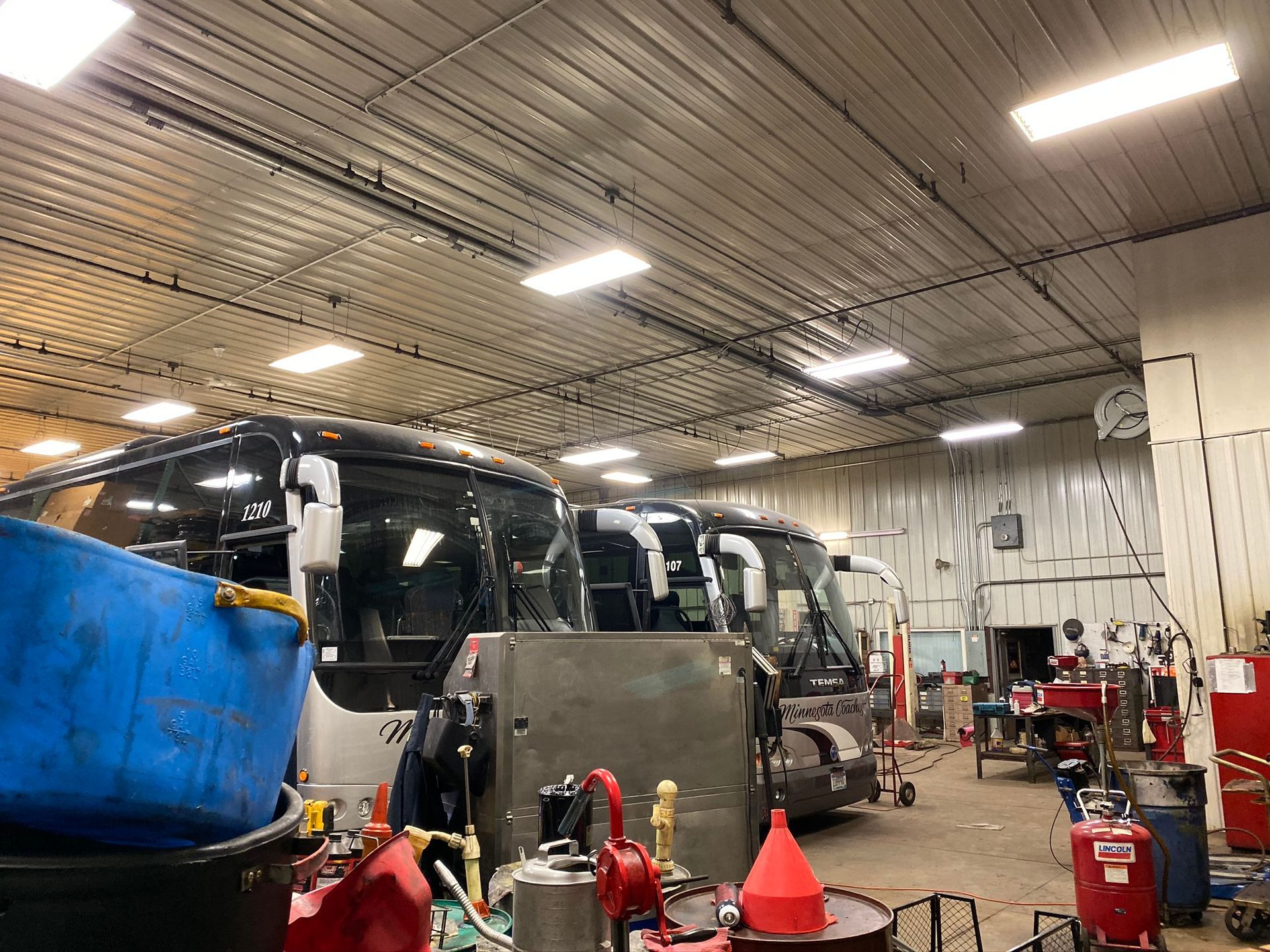 Buses inside a garage, next to tools and equipment. The interior has a metal ceiling with bright lights.