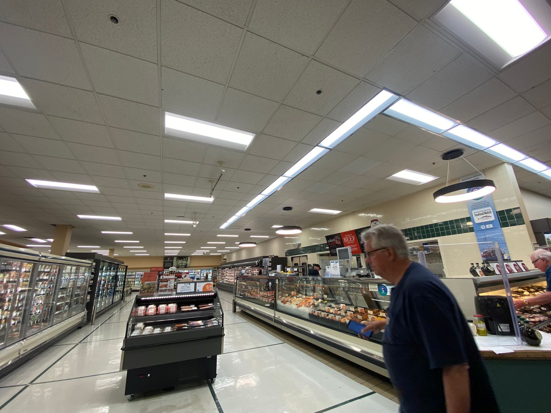 Grocery store interior; person browsing deli counter with various prepared food items.