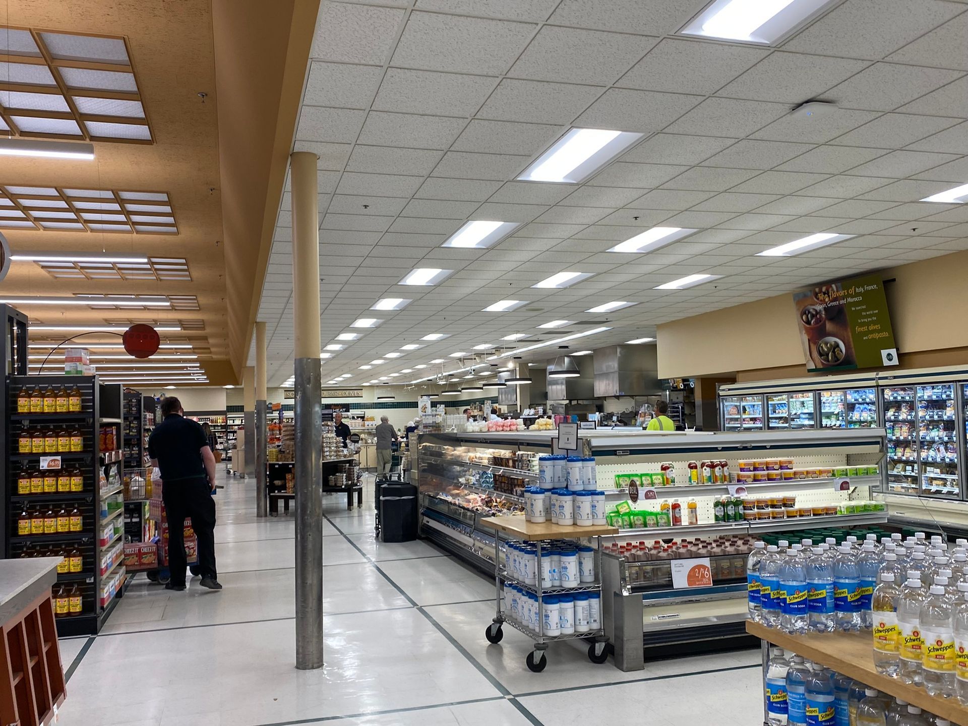 Grocery store interior with aisles, displays, and customers shopping. Brightly lit with overhead lights.