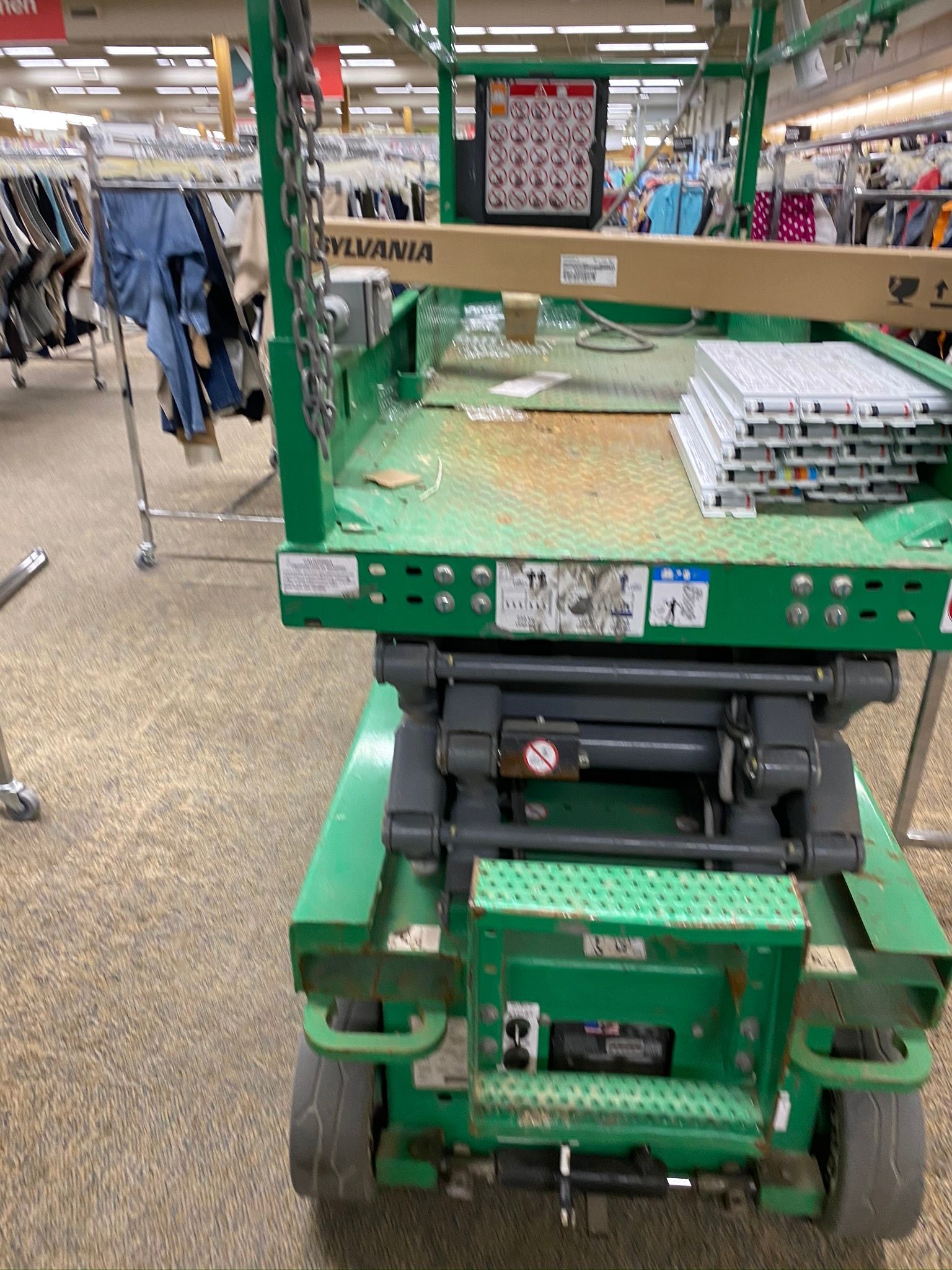 Green lift inside a retail store, with a stack of white shelving on the platform. Clothes racks and merchandise are visible in the background.