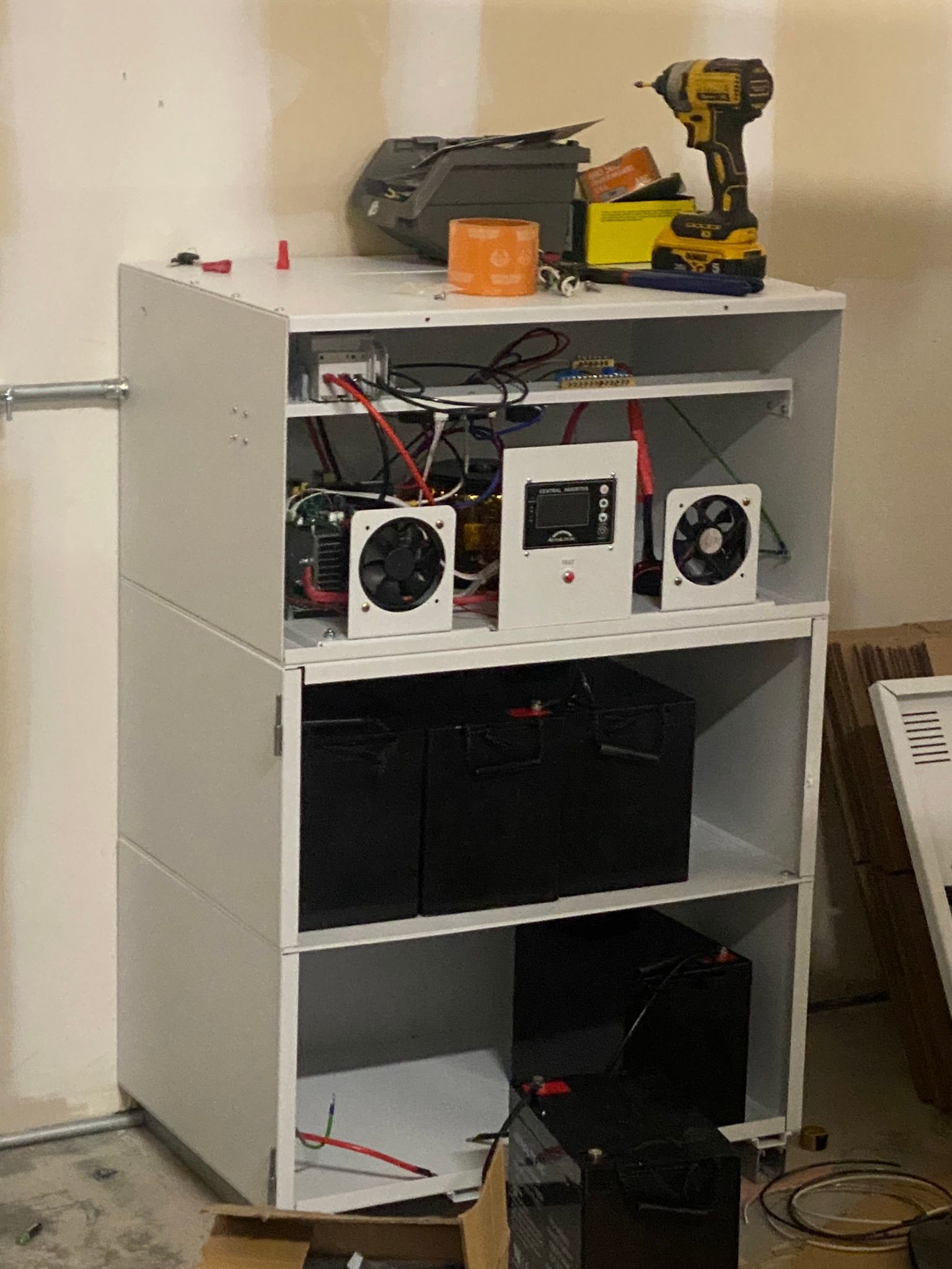 White shelving unit containing electrical components. Includes batteries, inverters, and fans. Tools on top shelf.
