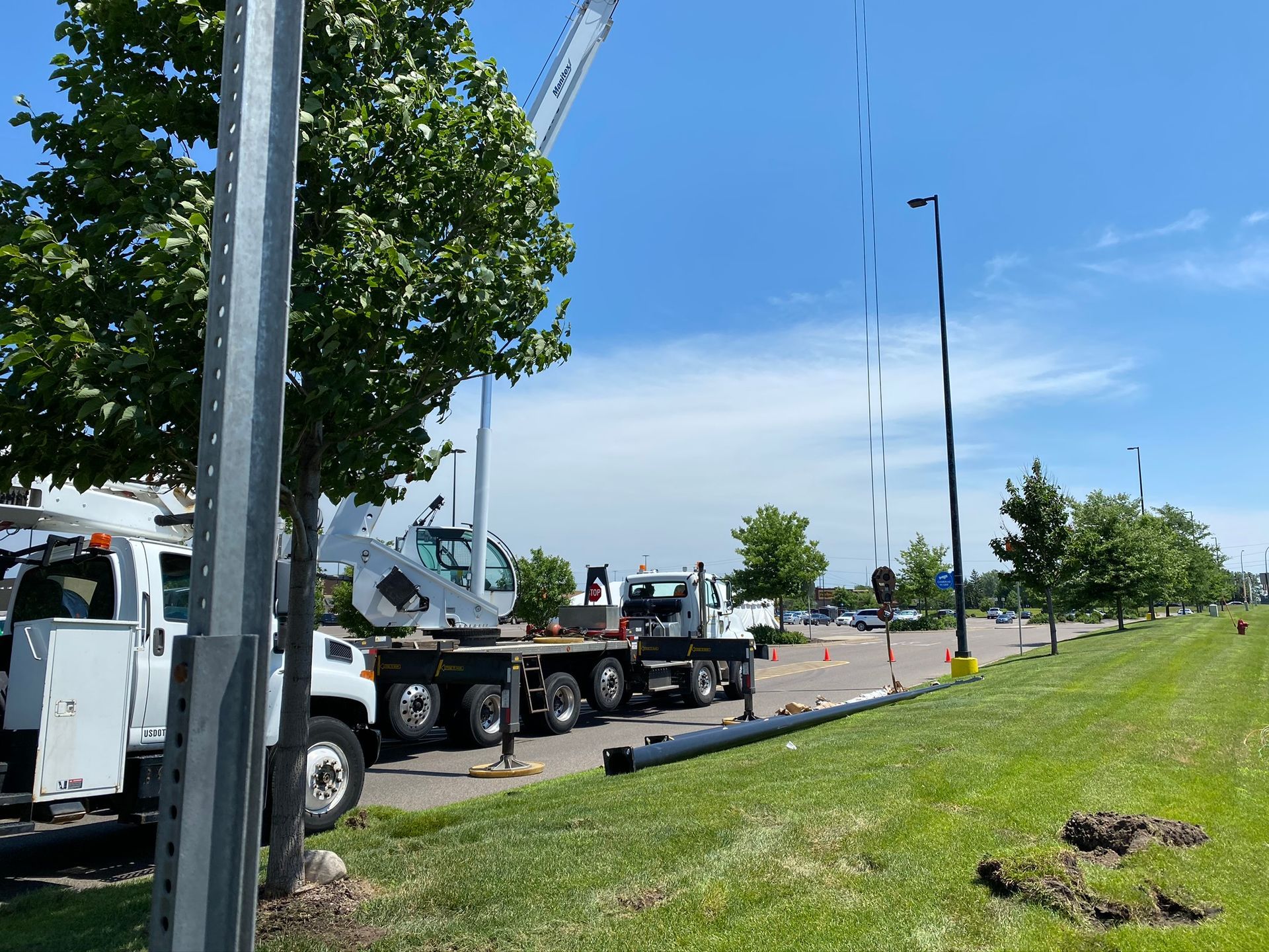 Construction trucks installing streetlights along a grassy median under a bright blue sky.