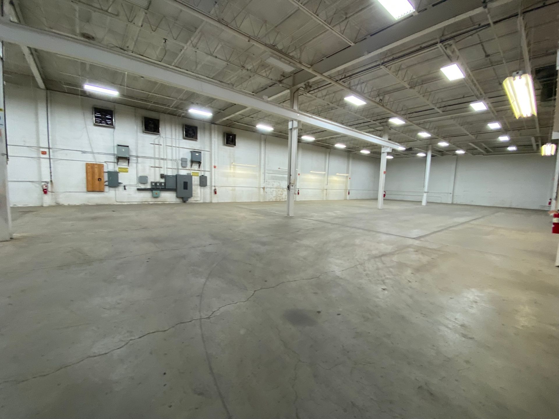 Empty, gray-floored warehouse interior with white walls, exposed beams, and overhead lighting.