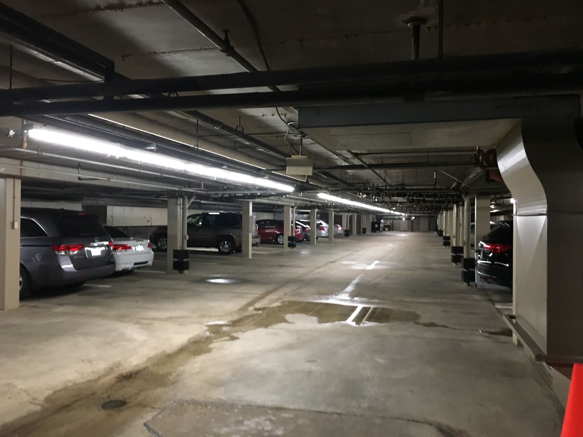 Parking garage with cars parked in stalls, fluorescent lights overhead, and puddles on the concrete floor.