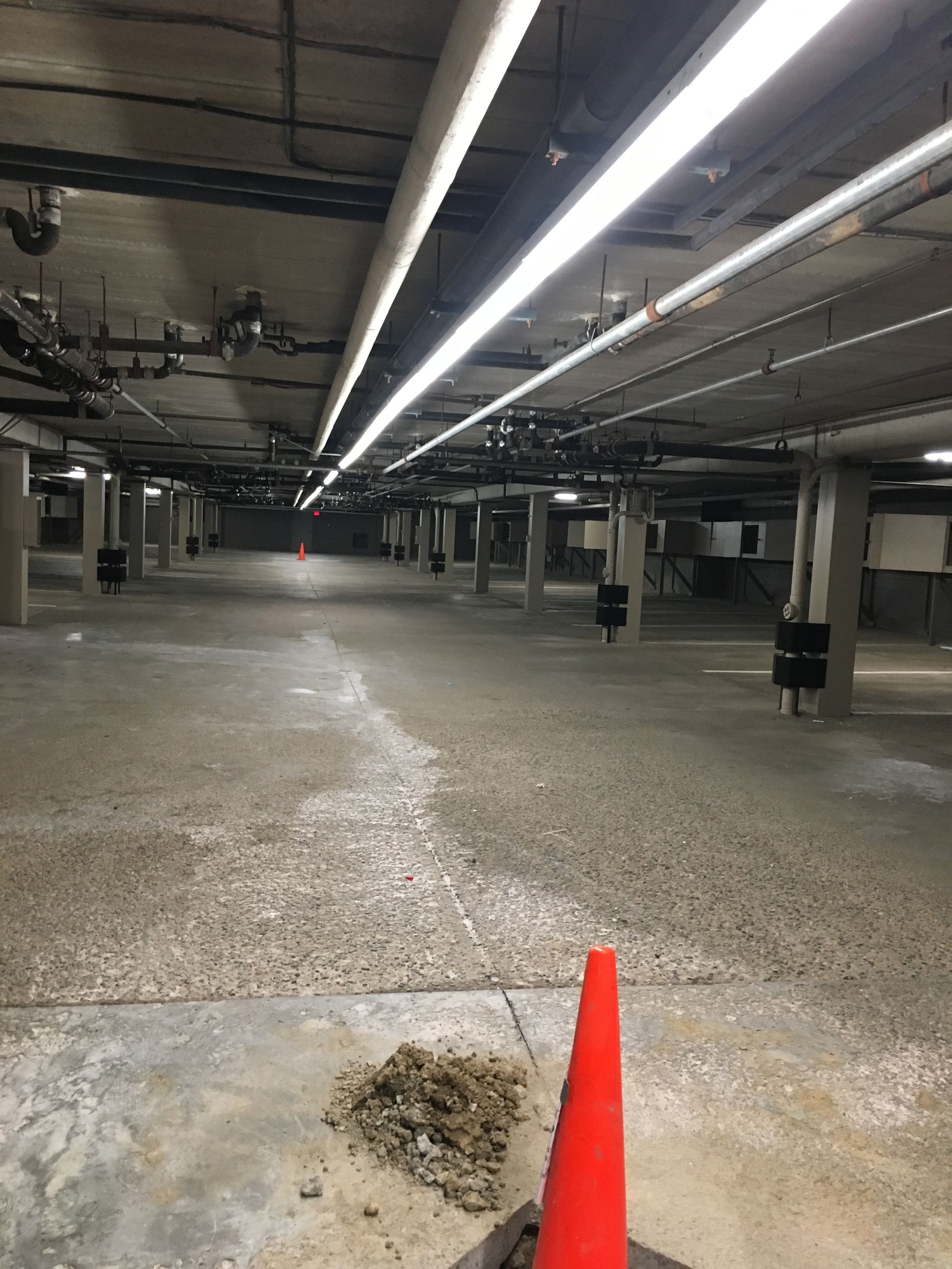 An empty concrete parking garage with overhead lights and columns. A traffic cone is in the foreground.