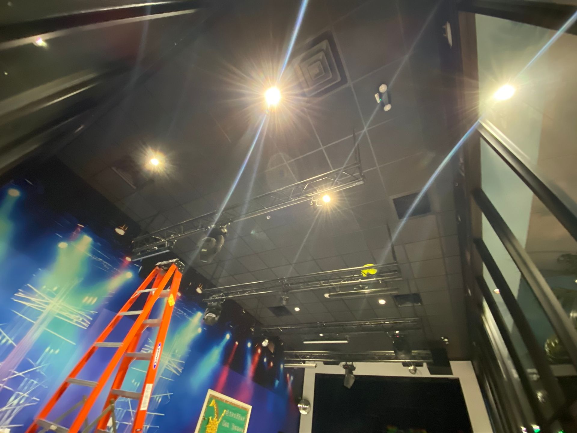 Stage with spotlights, ladder, and lighting rig. Blue stage backdrop, dark ceiling, and glass wall.