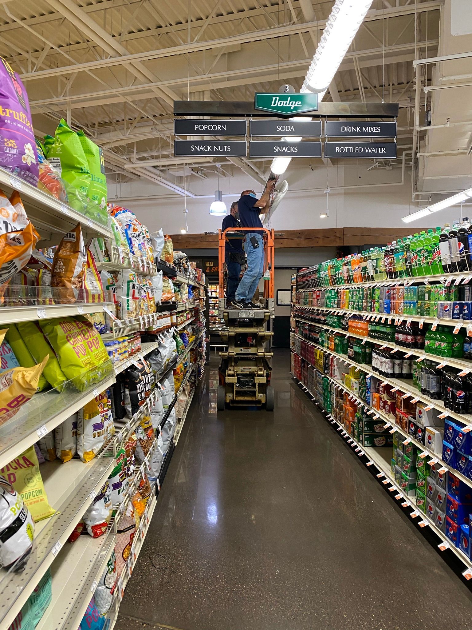 Person on a lift working in a grocery store aisle, surrounded by shelves of snacks and drinks.