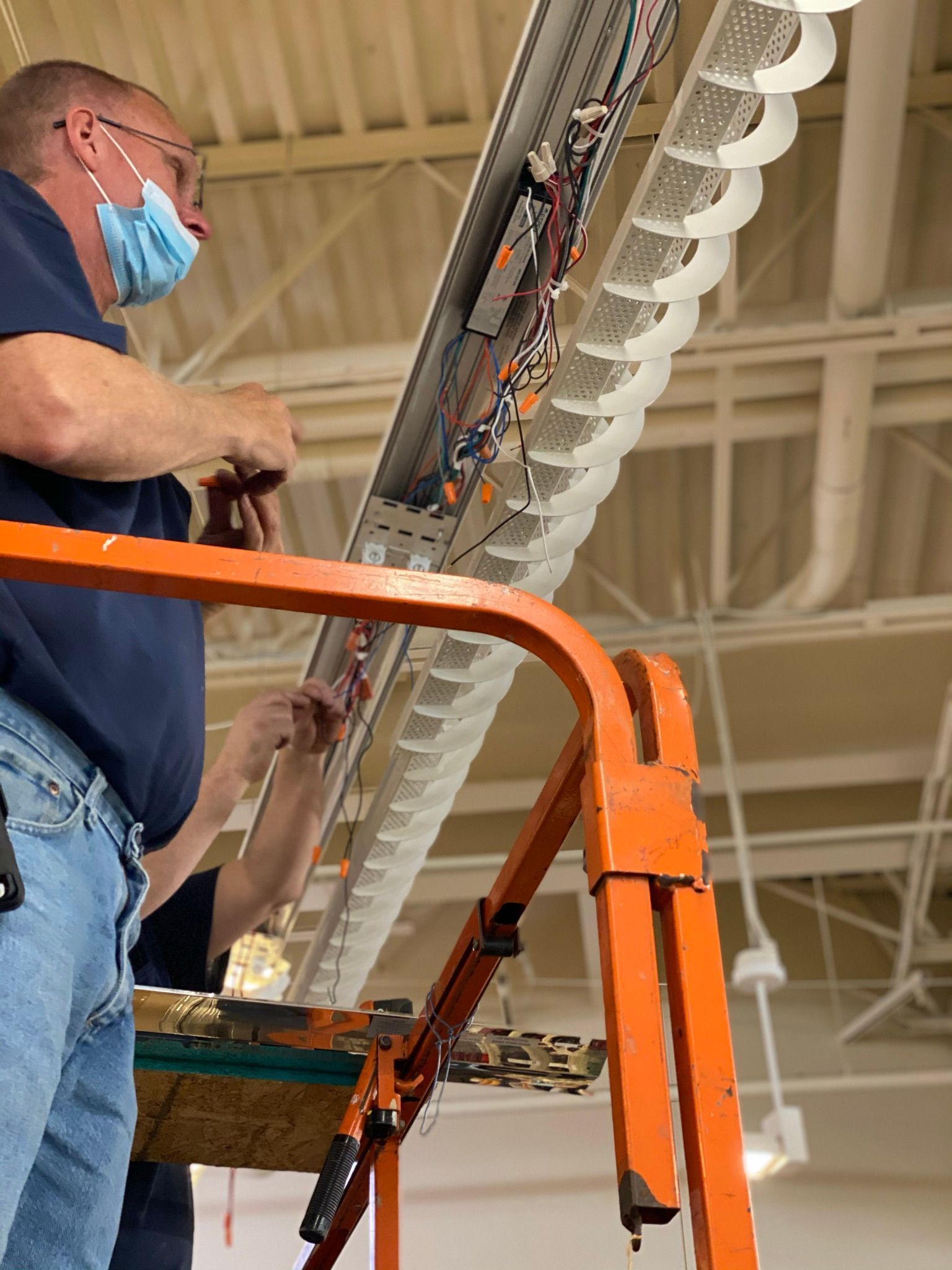 Man on lift, wearing mask, working on overhead lighting fixture with exposed wiring.