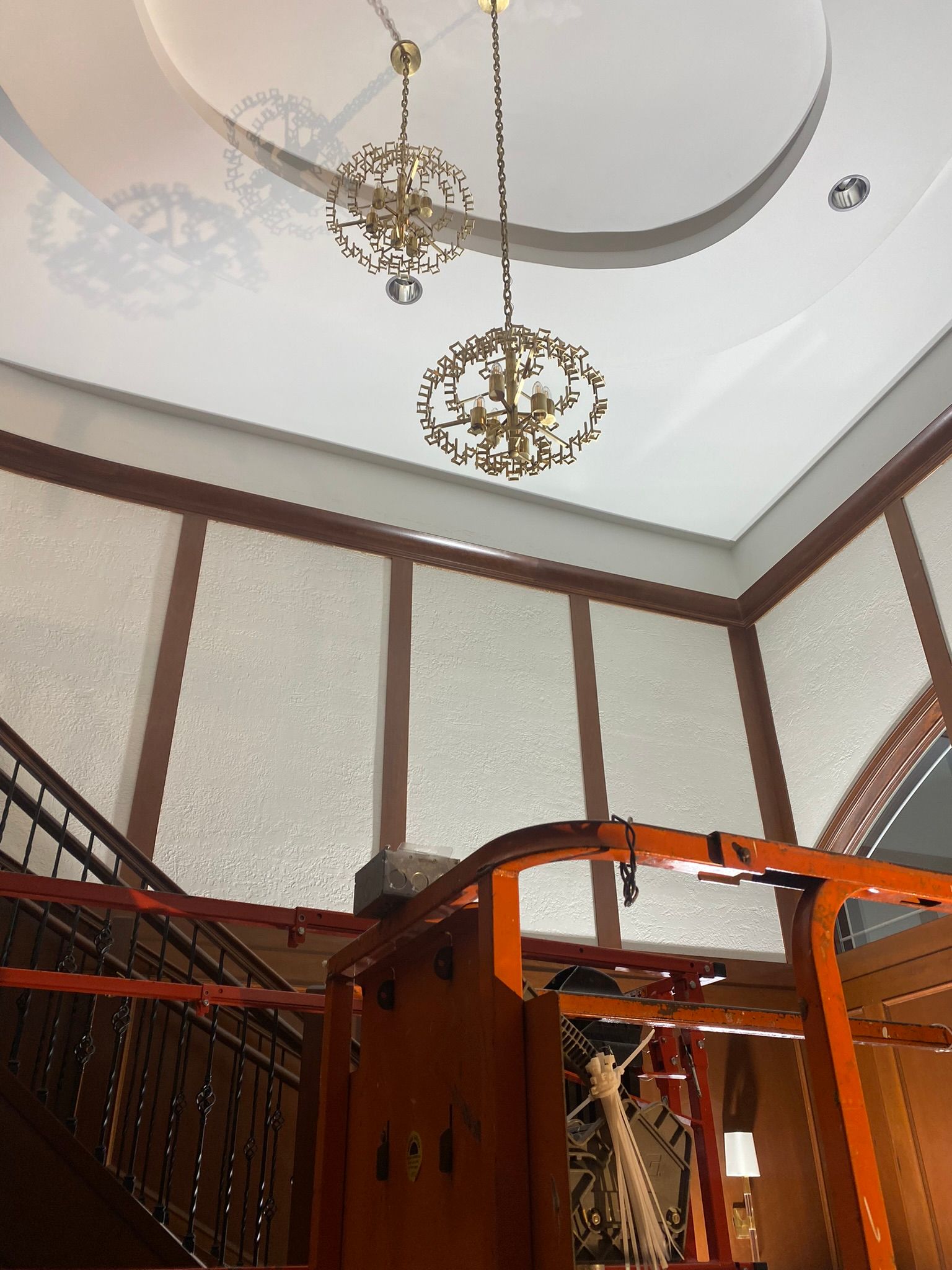 Two ornate chandeliers hang from a white ceiling with paneling and a dark staircase below.