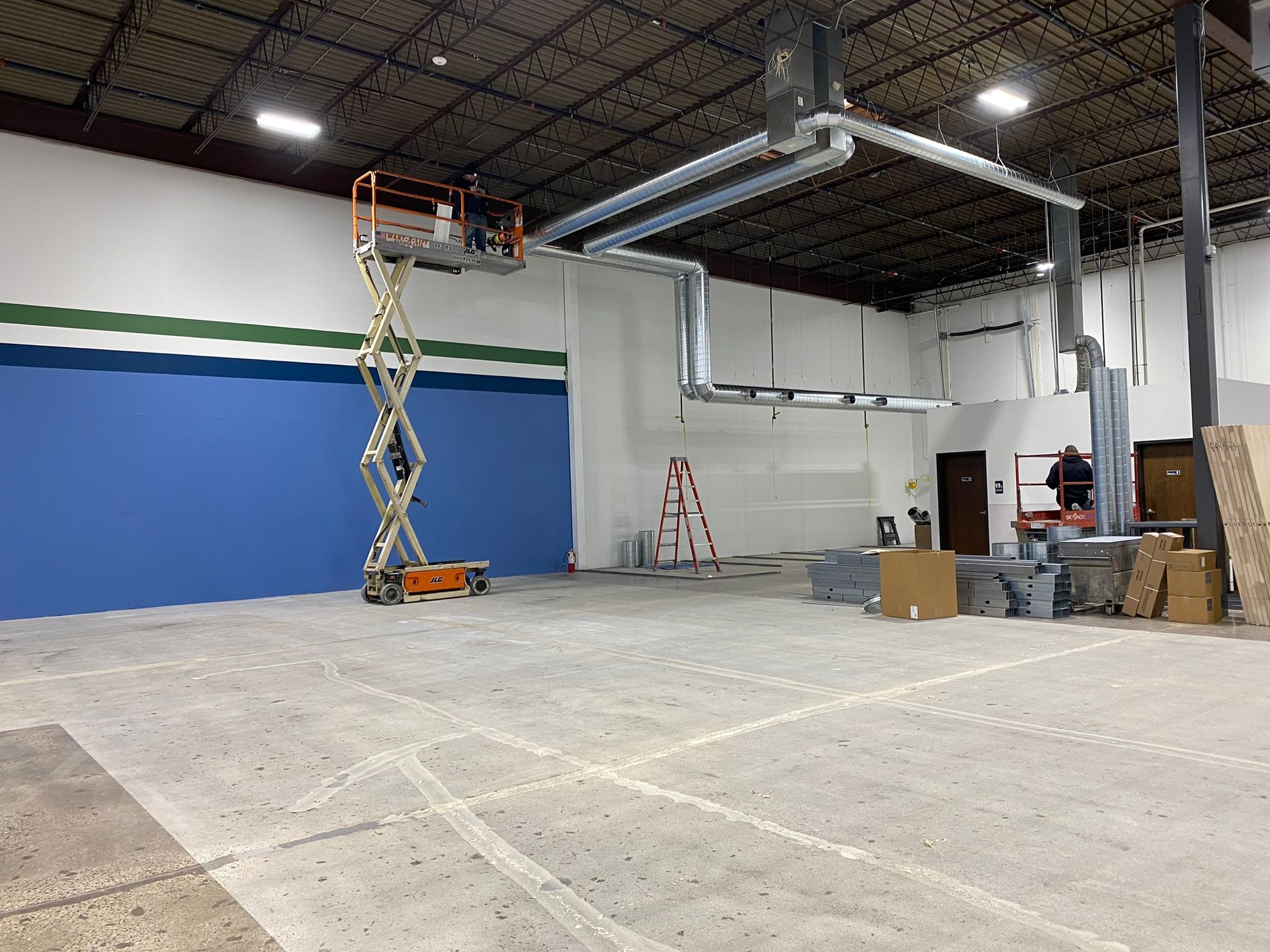 A person on a lift installs HVAC ductwork in a large, empty warehouse space.