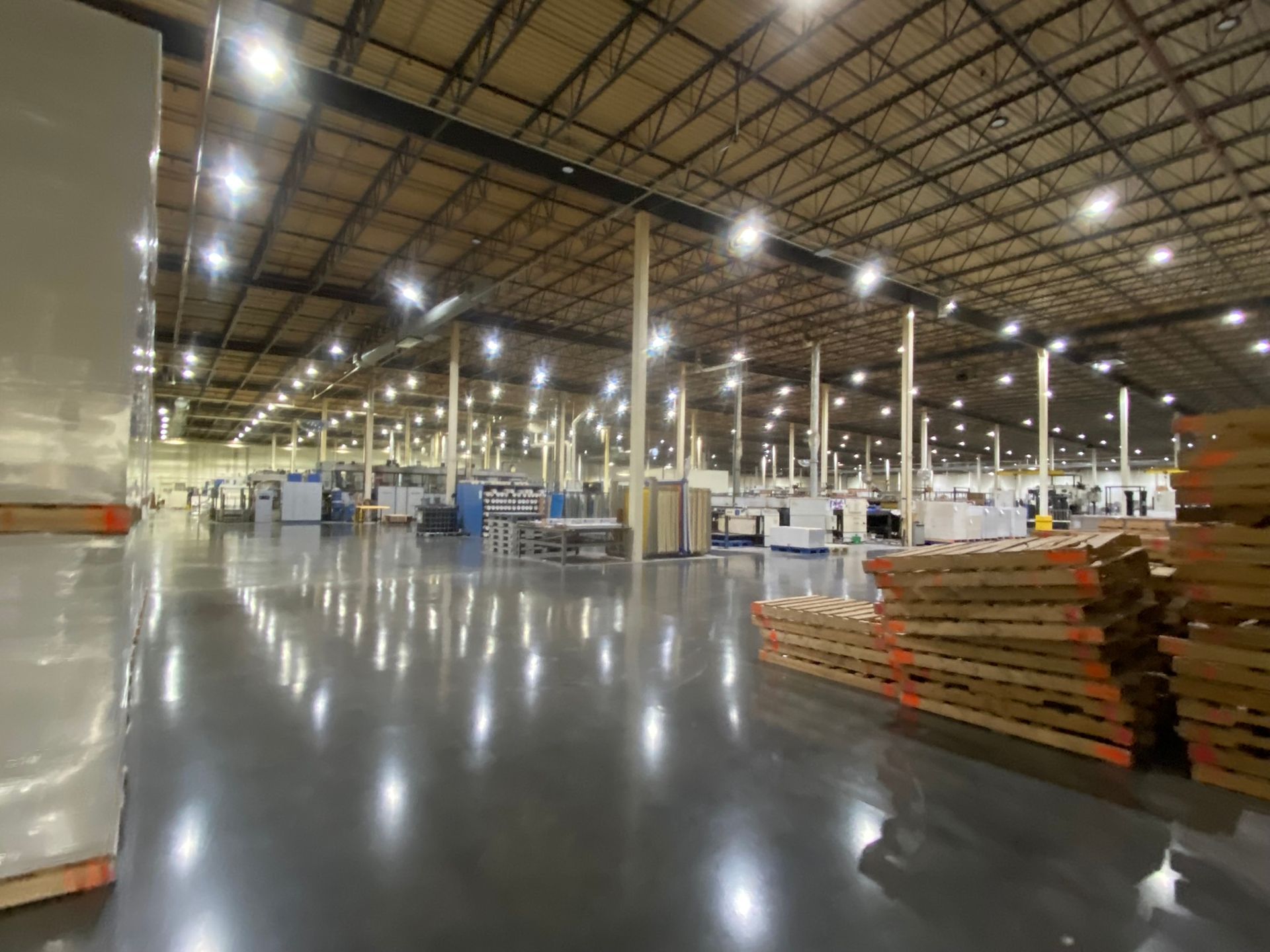 Large warehouse interior with industrial equipment and stacked pallets. Shiny floor reflects overhead lights.