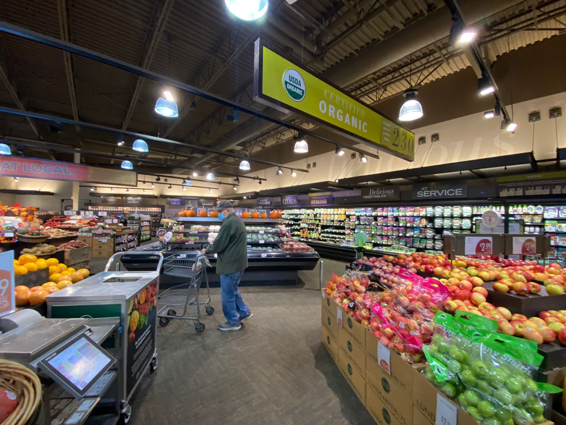 Grocery store interior with a person shopping in the produce section, organic signage overhead.