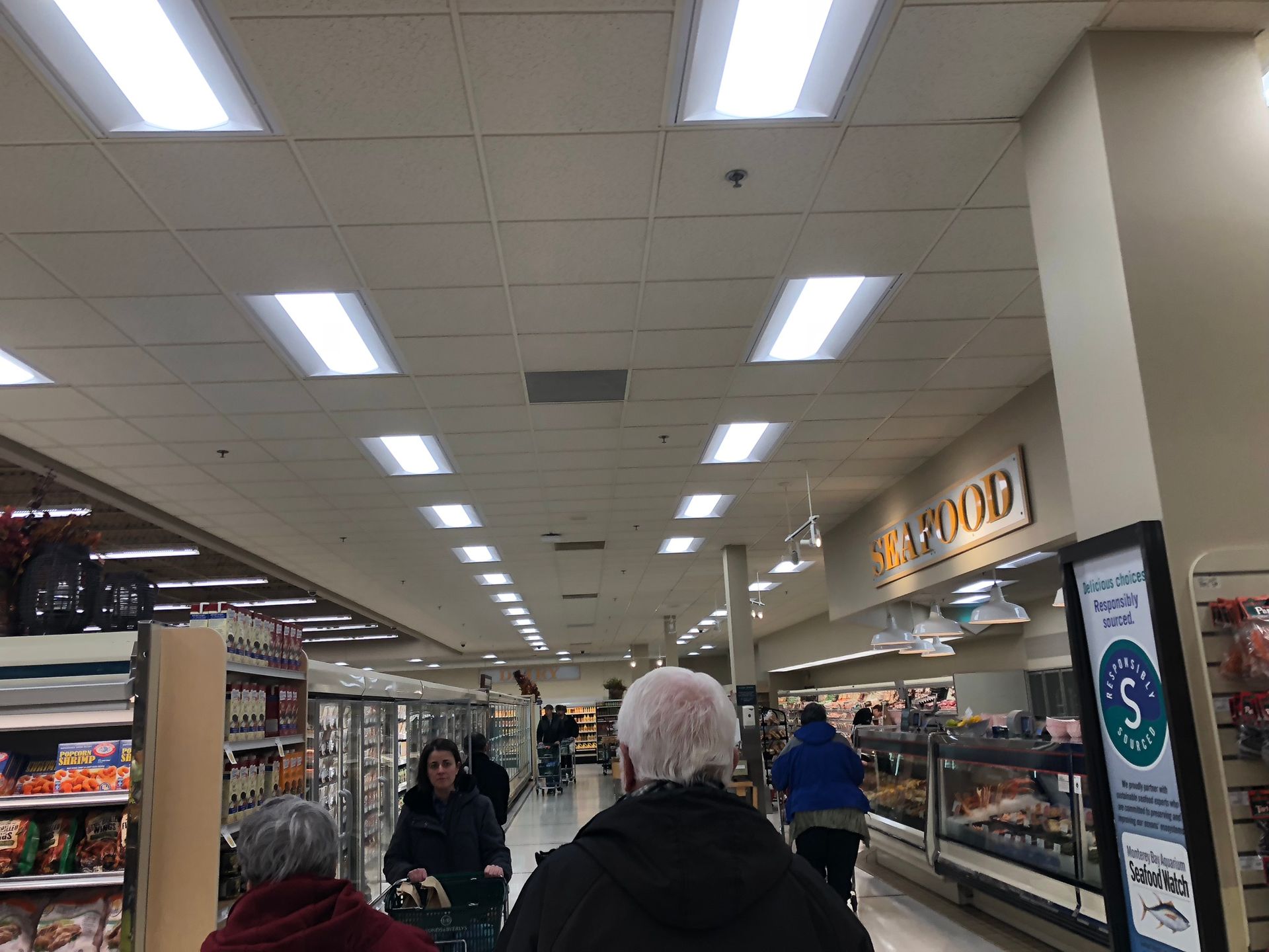 Grocery store aisle with overhead lighting. People with shopping carts are visible. 