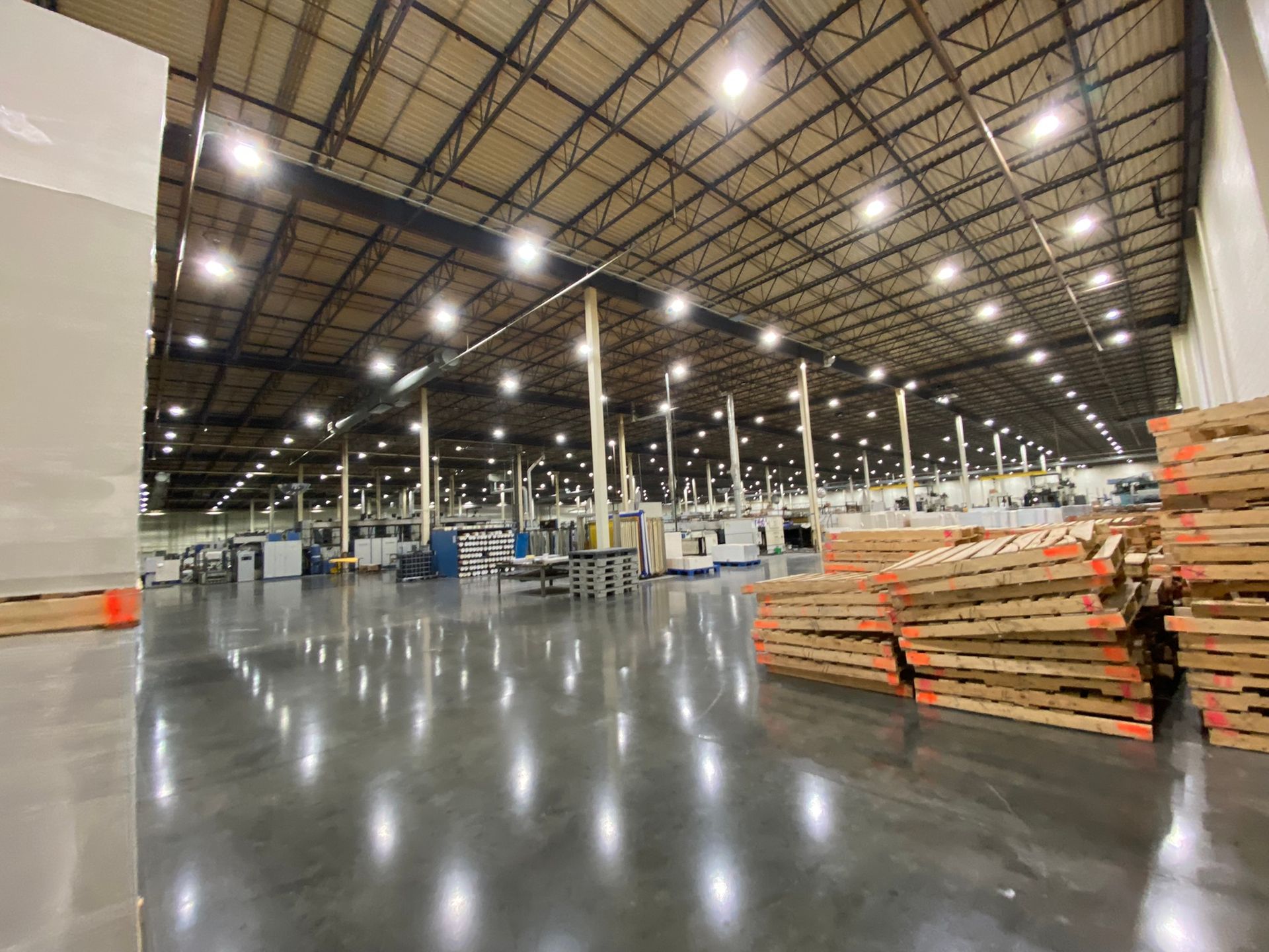 Large warehouse interior with industrial equipment and stacked wooden pallets on a reflective concrete floor.