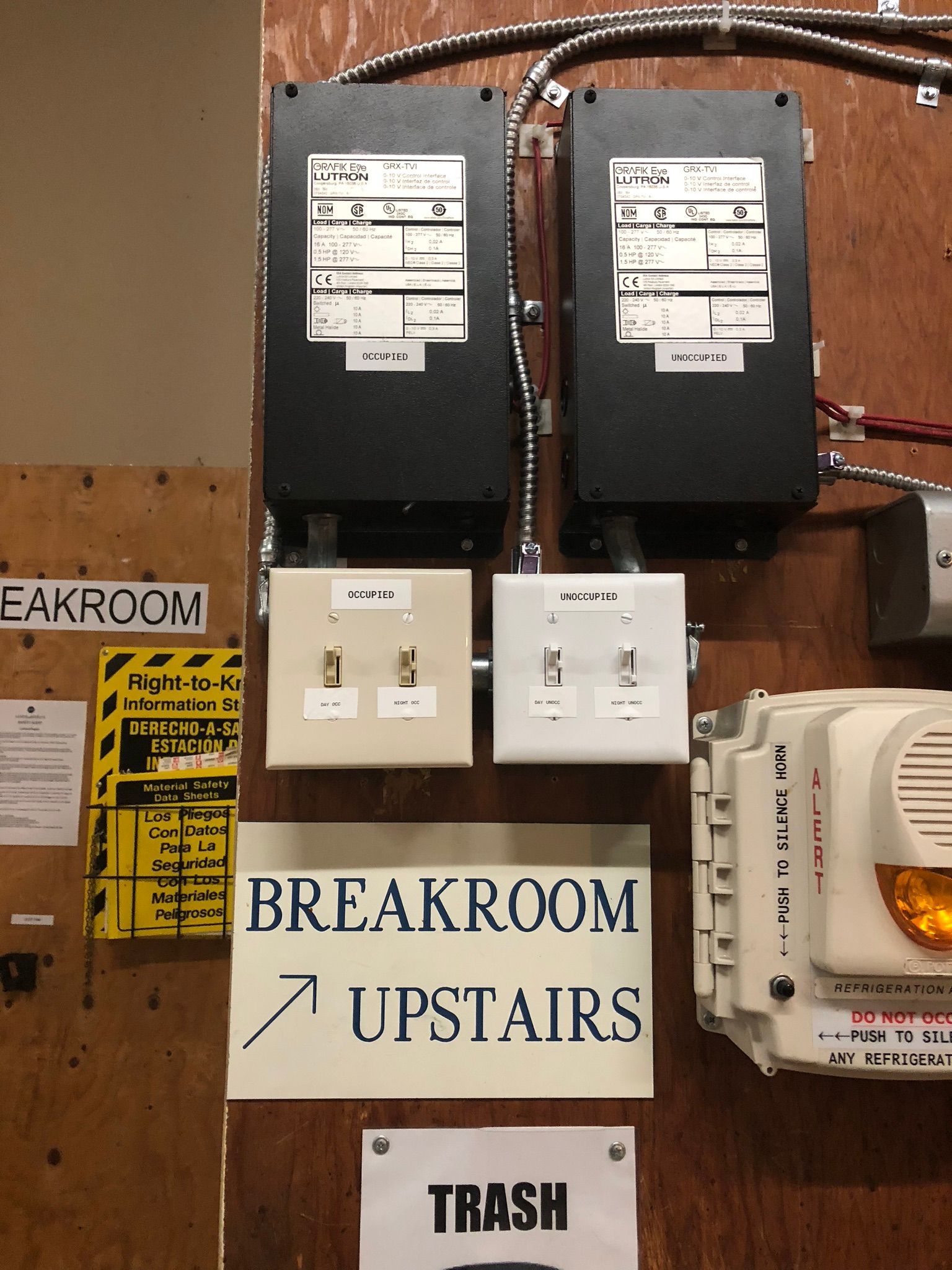 Two black boxes with labels and outlets, a breakroom sign, and a trash sign mounted on a wooden wall.