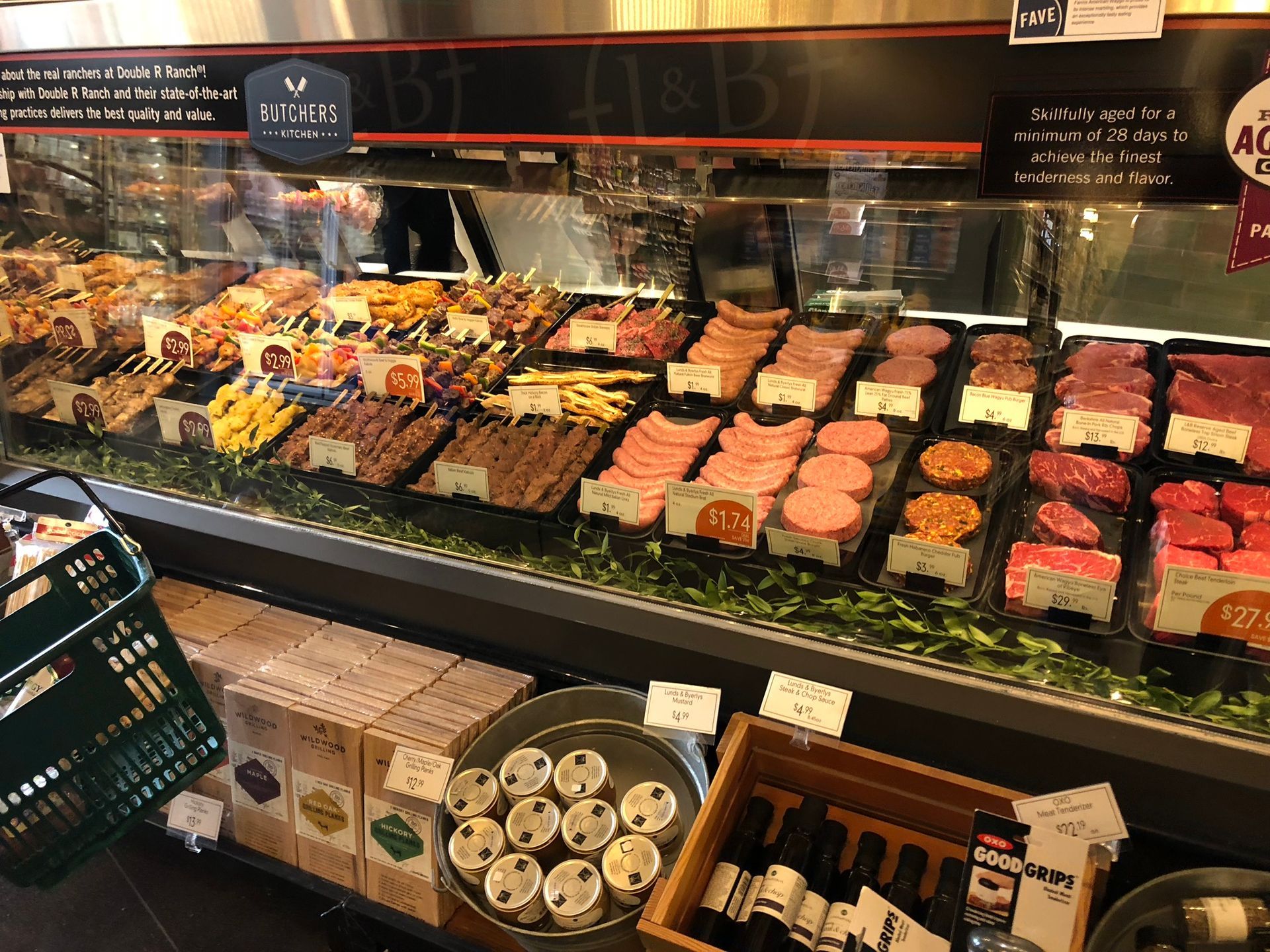 Meat display case in a store, filled with various prepared foods. Green basket and products are in front.