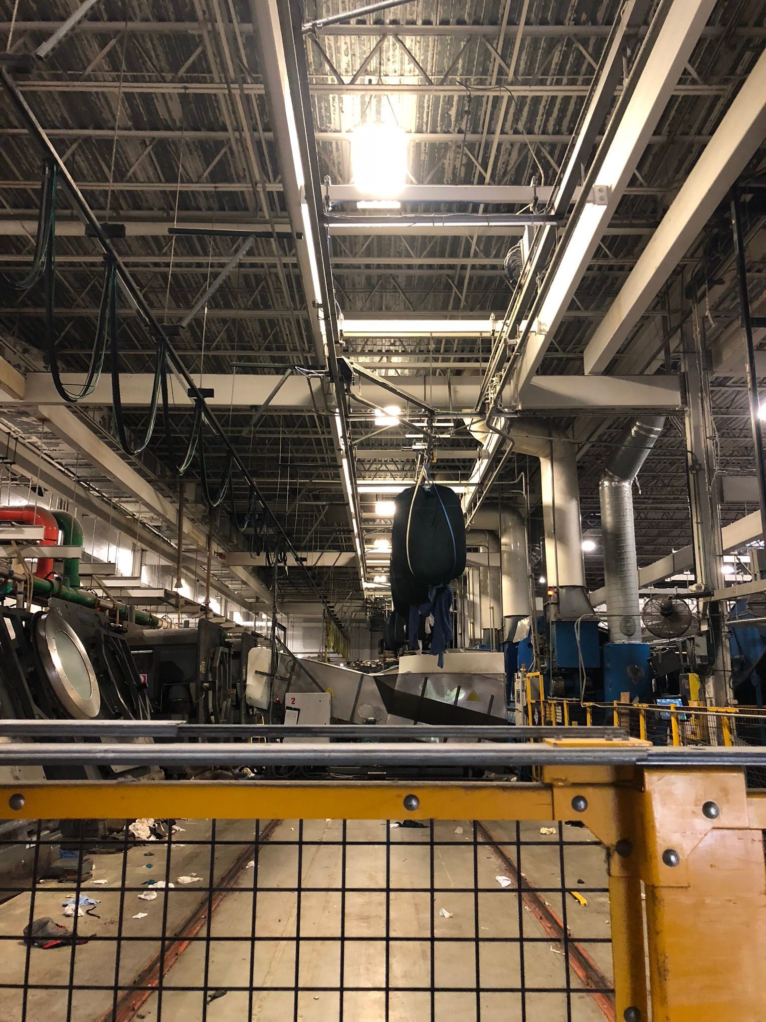 Factory interior with overhead conveyor system and machinery. Yellow barrier in foreground.