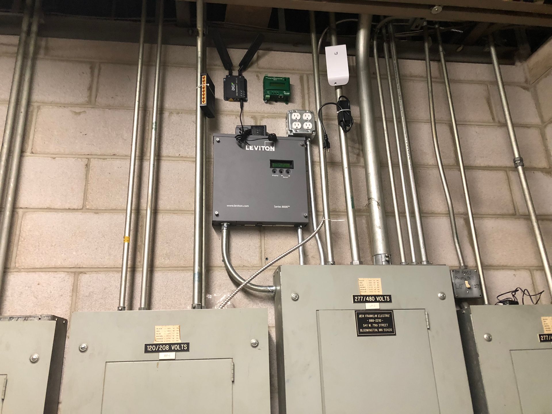 Electrical equipment mounted on a brick wall, including conduit, circuit breaker boxes, and electronic devices.