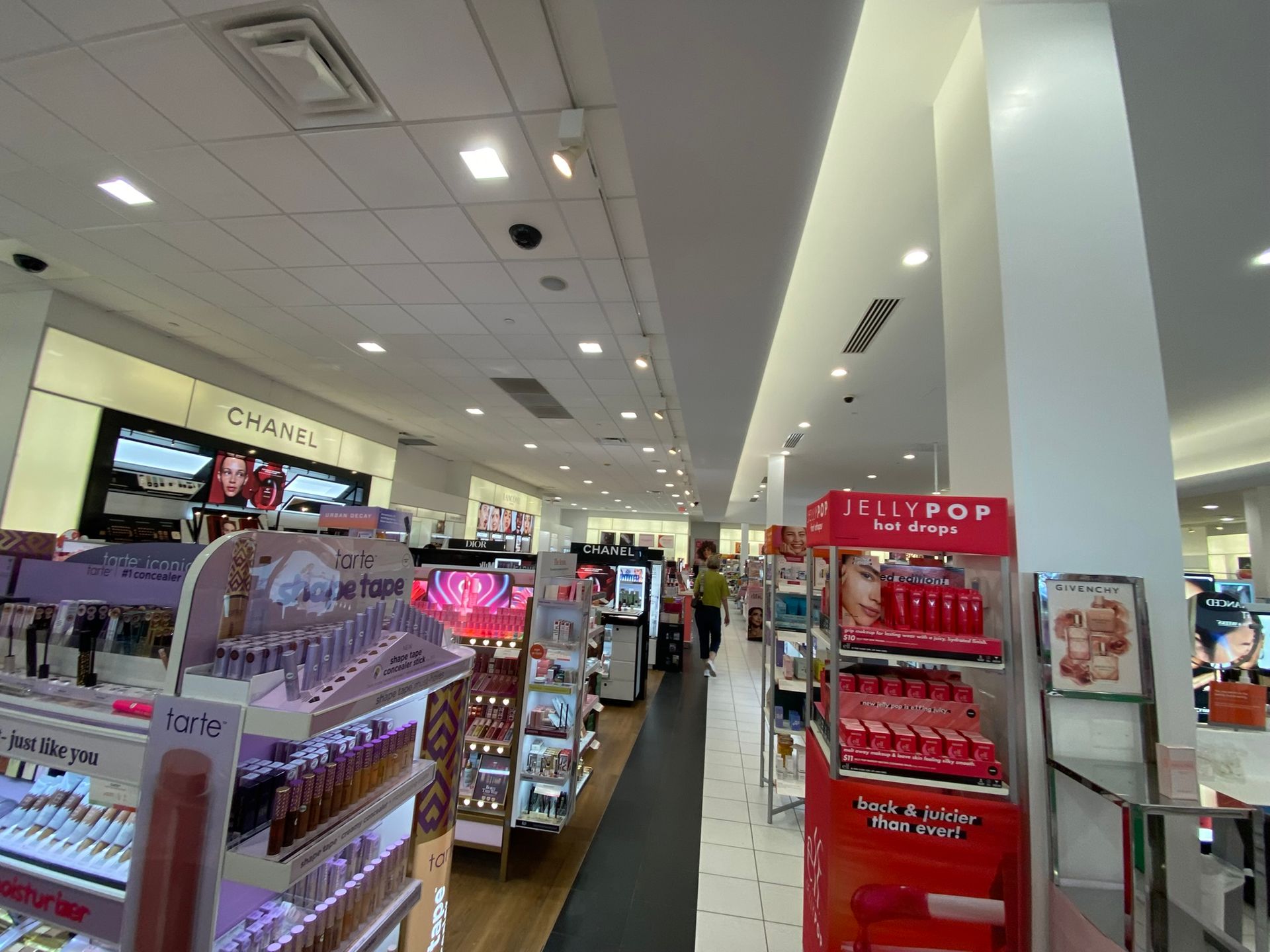 Cosmetics aisle in a department store with various makeup displays and shoppers. Bright lighting.