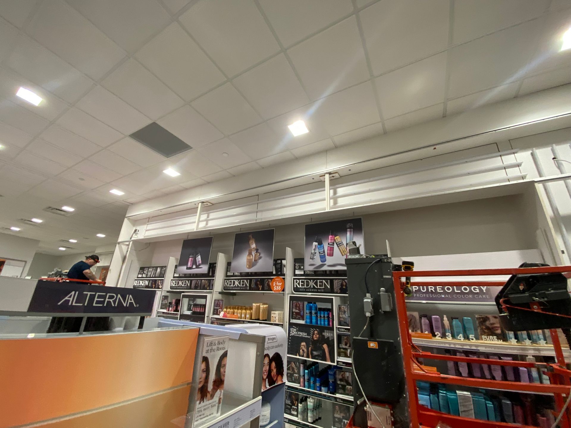 Inside a store, hair product display. Shelves and posters, a person in the background. Bright lighting.