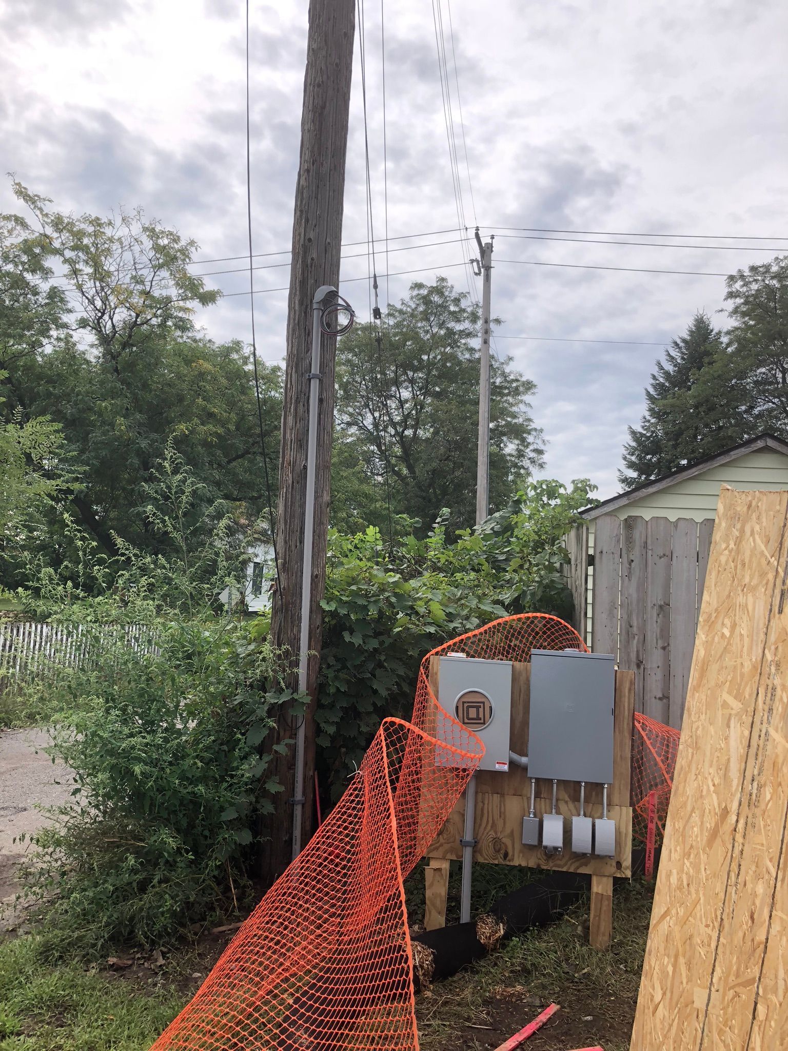 Wooden utility pole with electrical box, orange safety fence, and construction materials in a grassy area.