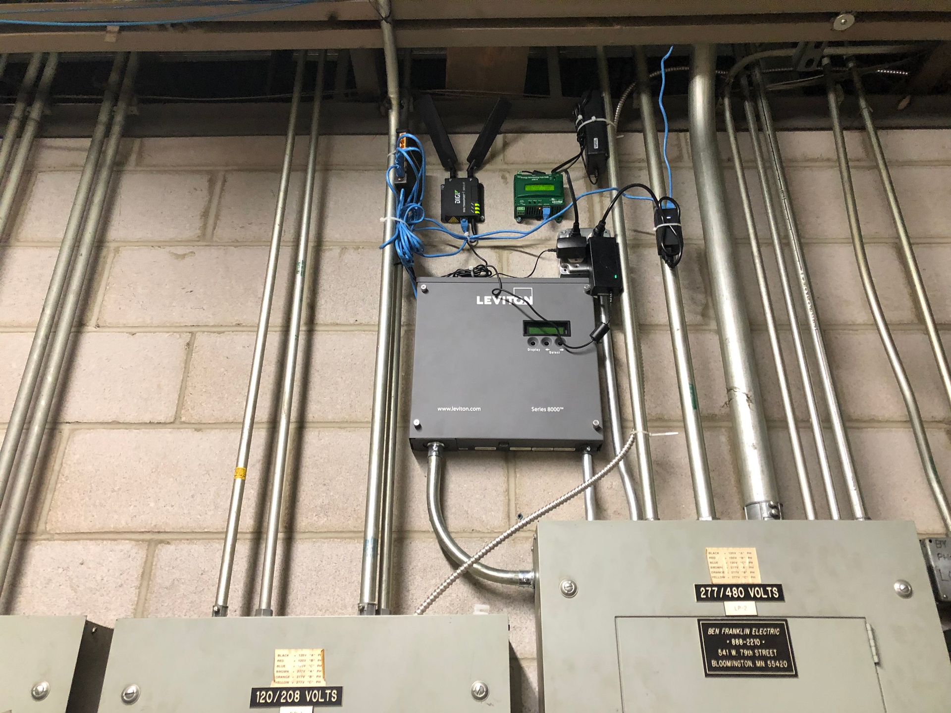 Electrical equipment mounted on a brick wall, including conduit, a grey box, and smaller black and green devices.
