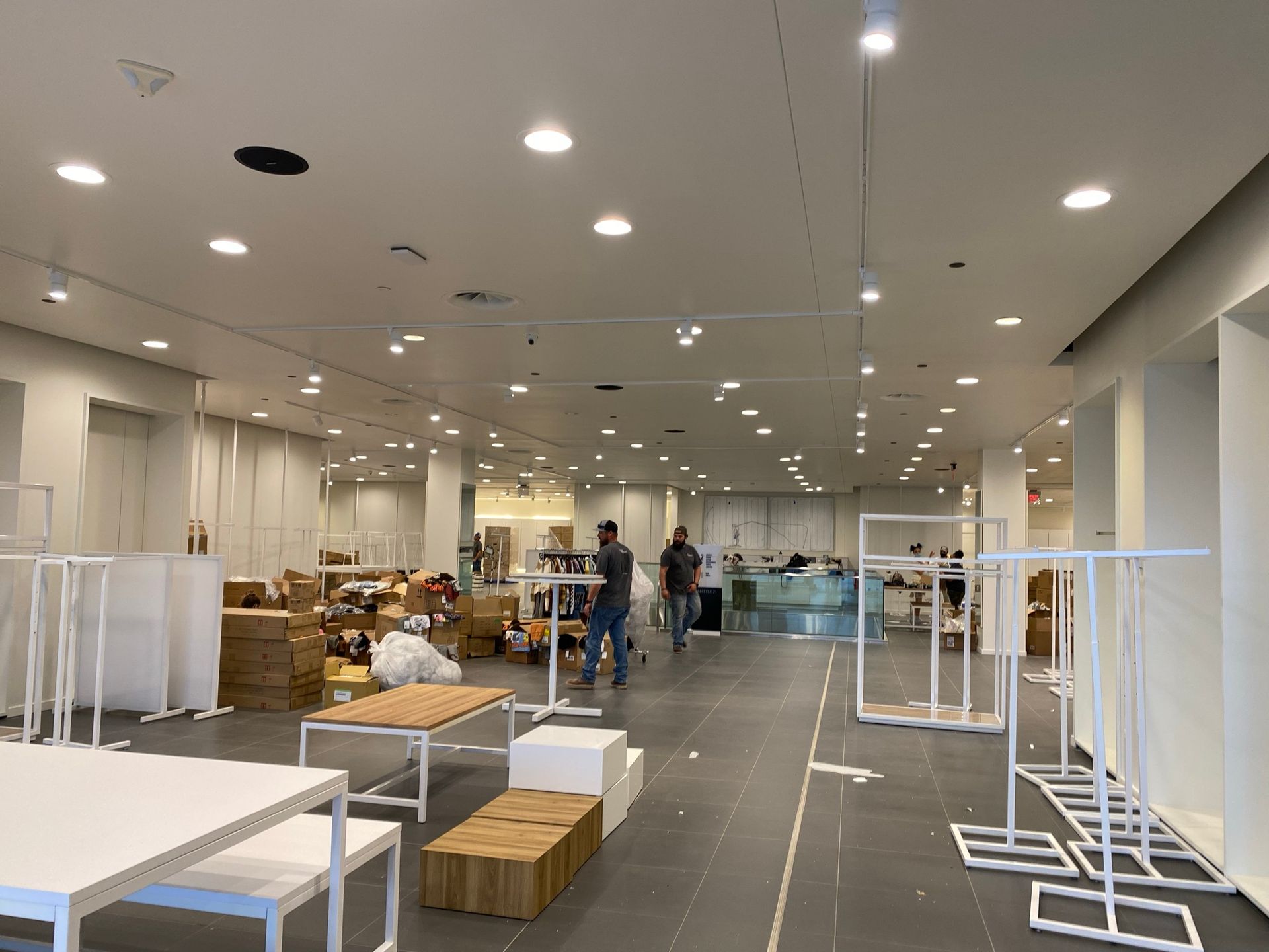 Empty retail space with workers. White walls, display tables, clothing racks, boxes, and recessed lighting.