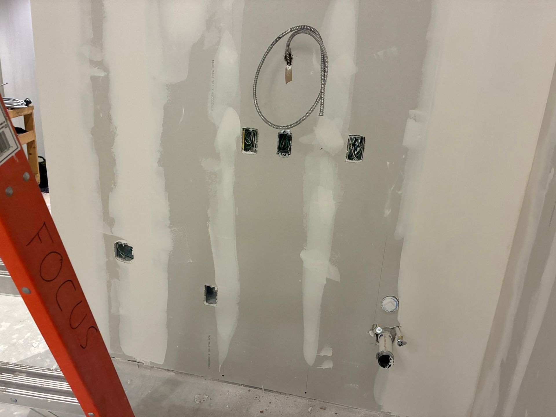 A wall with drywall, electrical boxes, and wiring, in a construction setting. An orange ladder is in the frame.