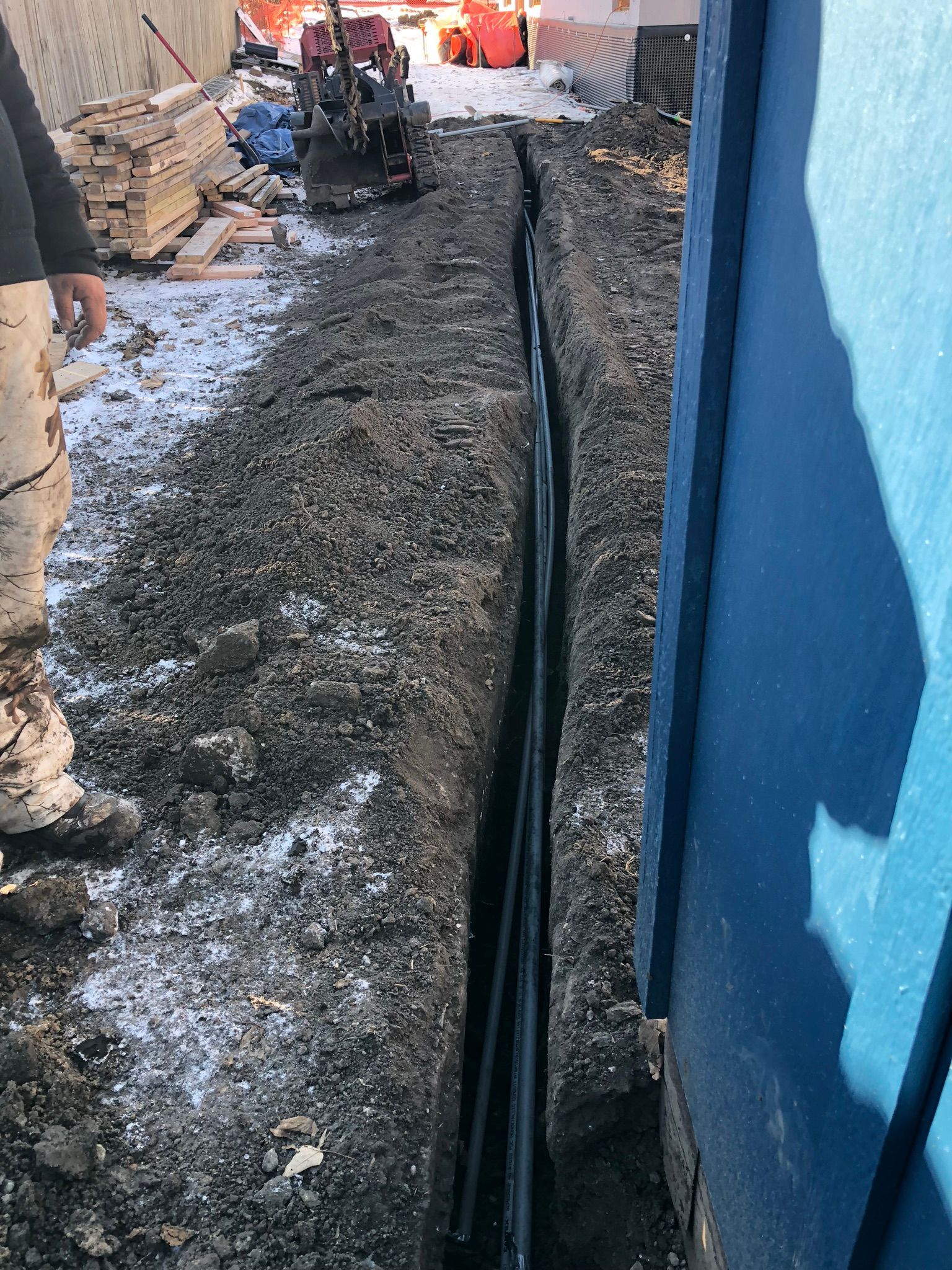 Trench with black pipes along a blue building. A small excavator and person's legs are visible near dirt and snow.