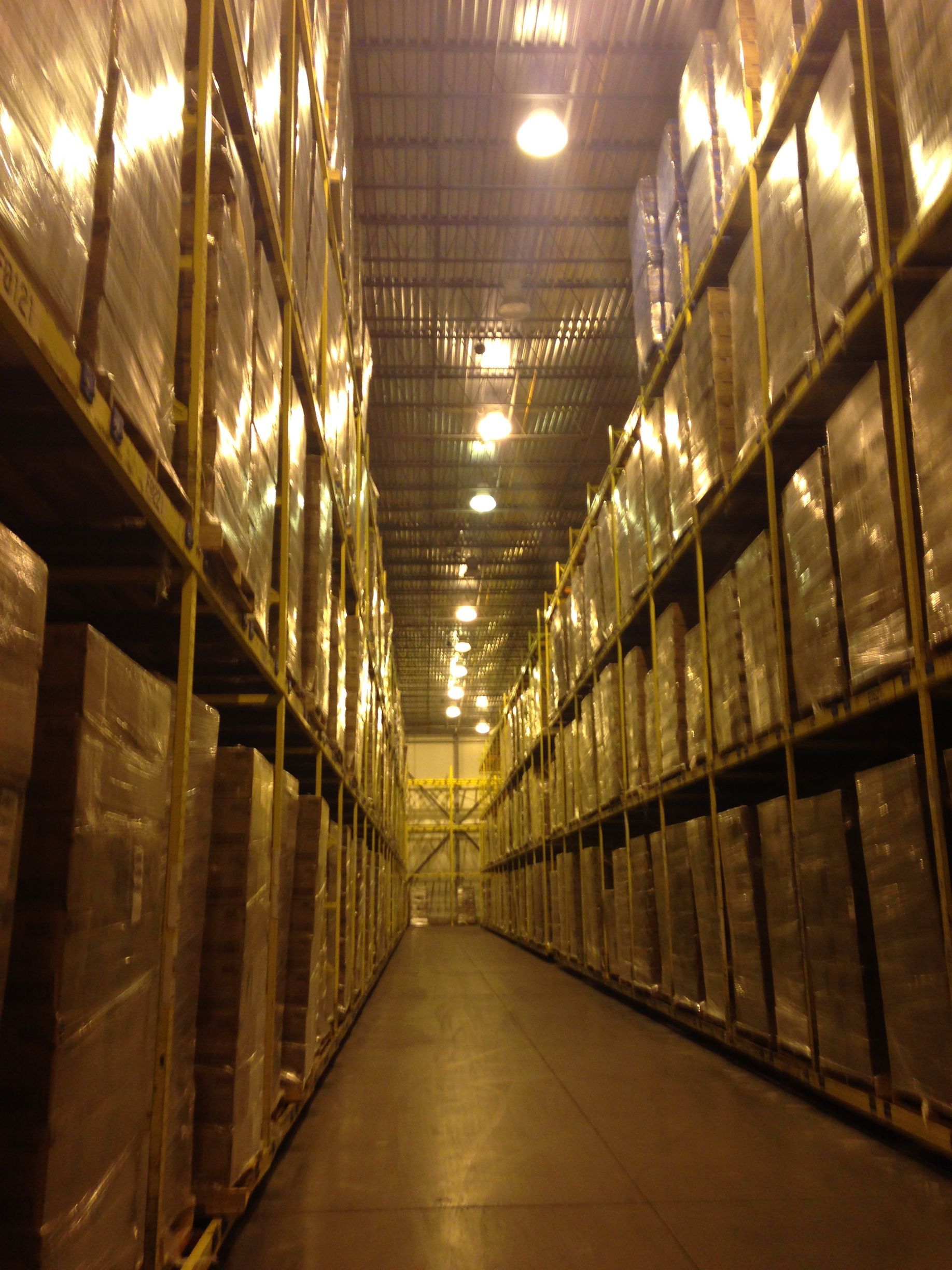 Warehouse aisle with tall racks of wrapped boxes, lit by overhead lights.