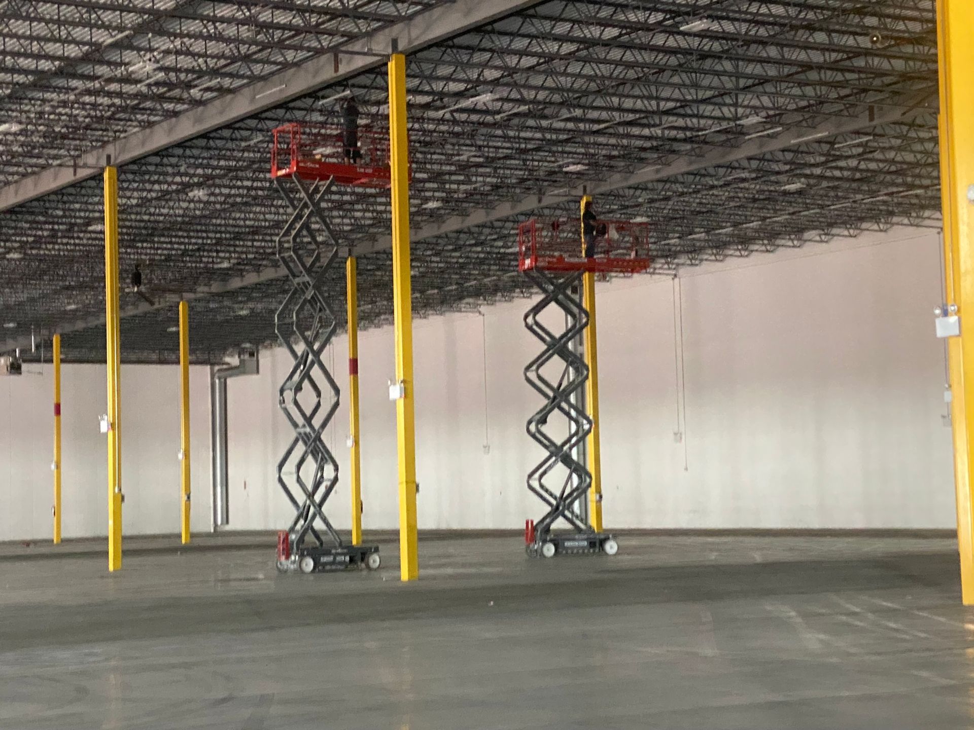 Two scissor lifts inside a large warehouse, near yellow support beams, with workers on the lifts.