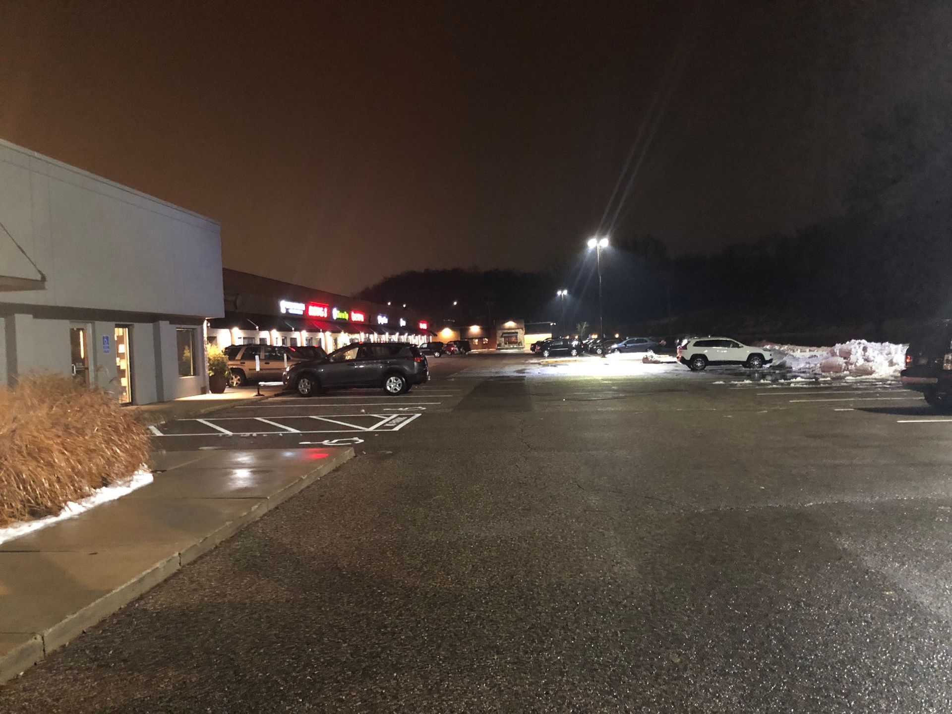 Nighttime view of a strip mall with cars parked in front of various businesses, wet asphalt, dark sky.