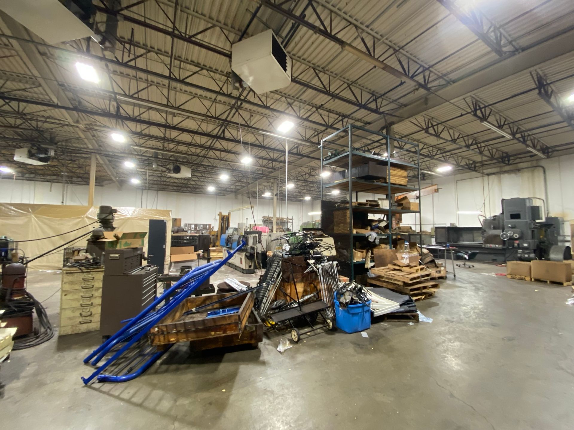 Warehouse interior, cluttered with equipment, shelving, and pallets. High ceilings, overhead lighting, and concrete floor.