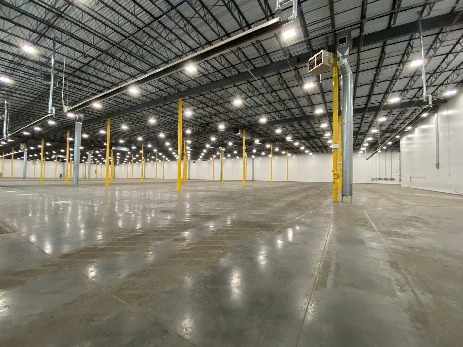 Large empty warehouse interior with a gray concrete floor, yellow support beams, and bright overhead lights.
