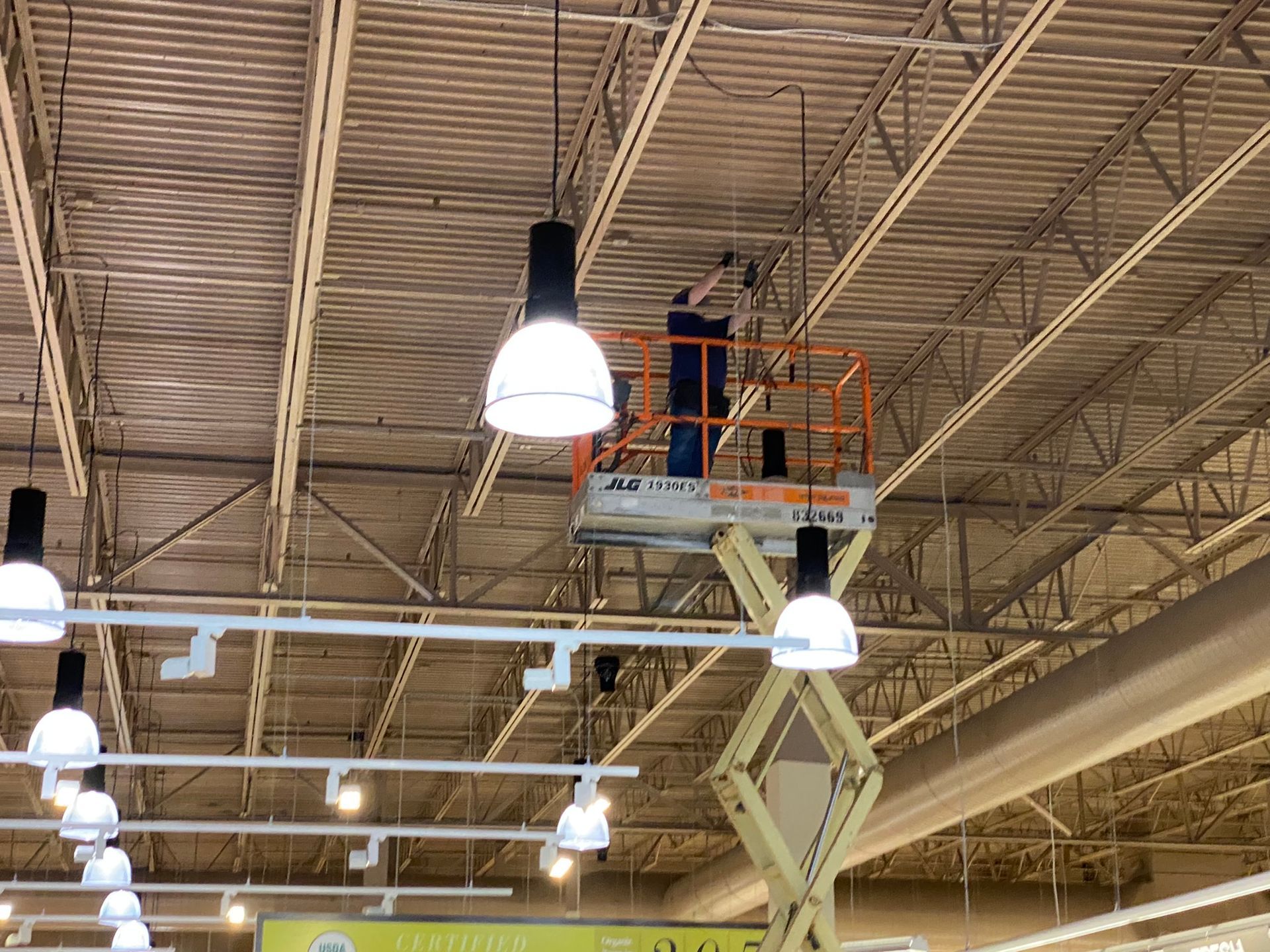 Person on lift repairs overhead light in a commercial building with white ceiling and steel beams.
