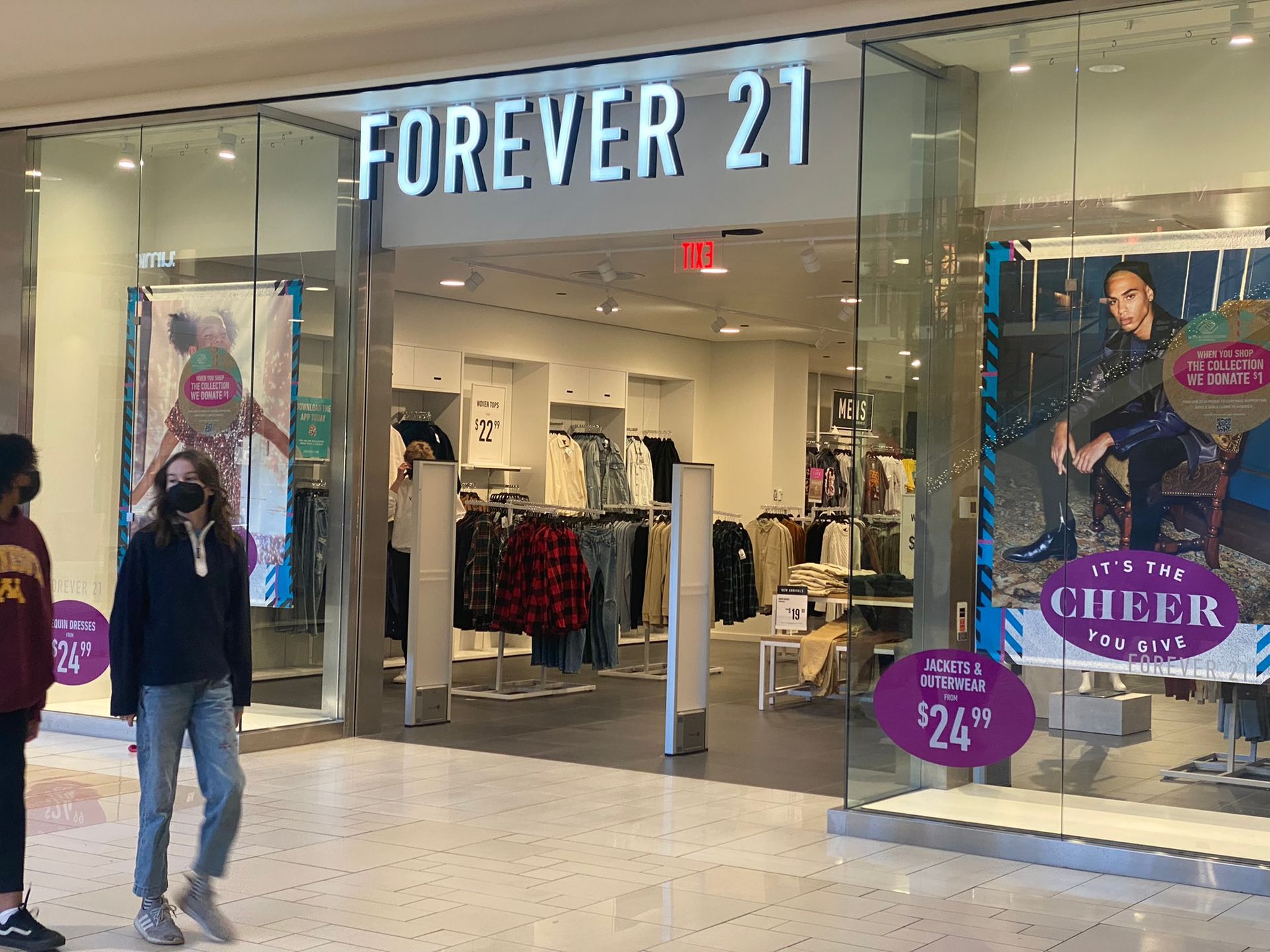 Forever 21 store entrance. A person in a mask walks past glass doors. Clothing racks inside.