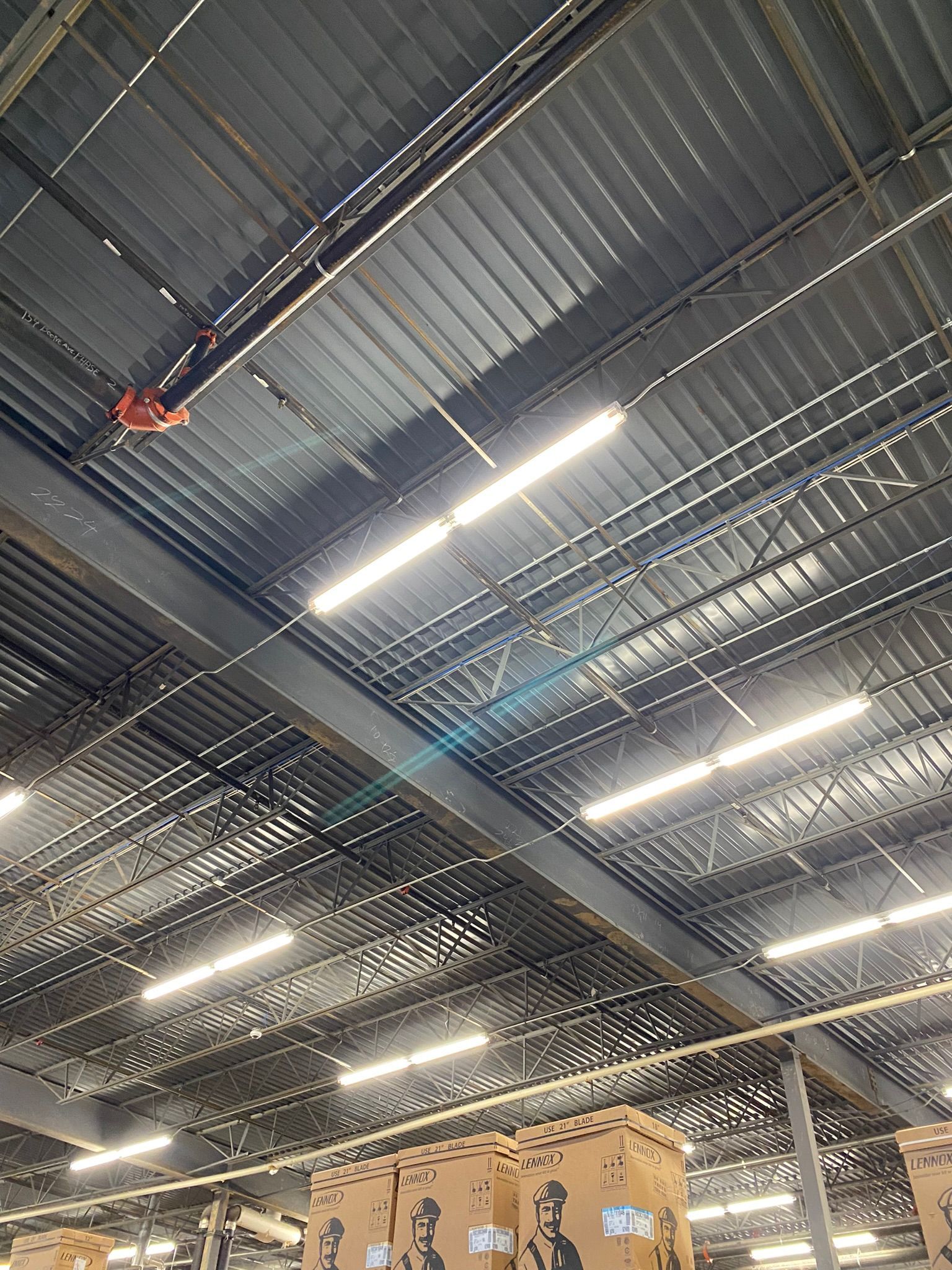 Warehouse ceiling with metal beams and corrugated panels, bright fluorescent lights, and boxes below.