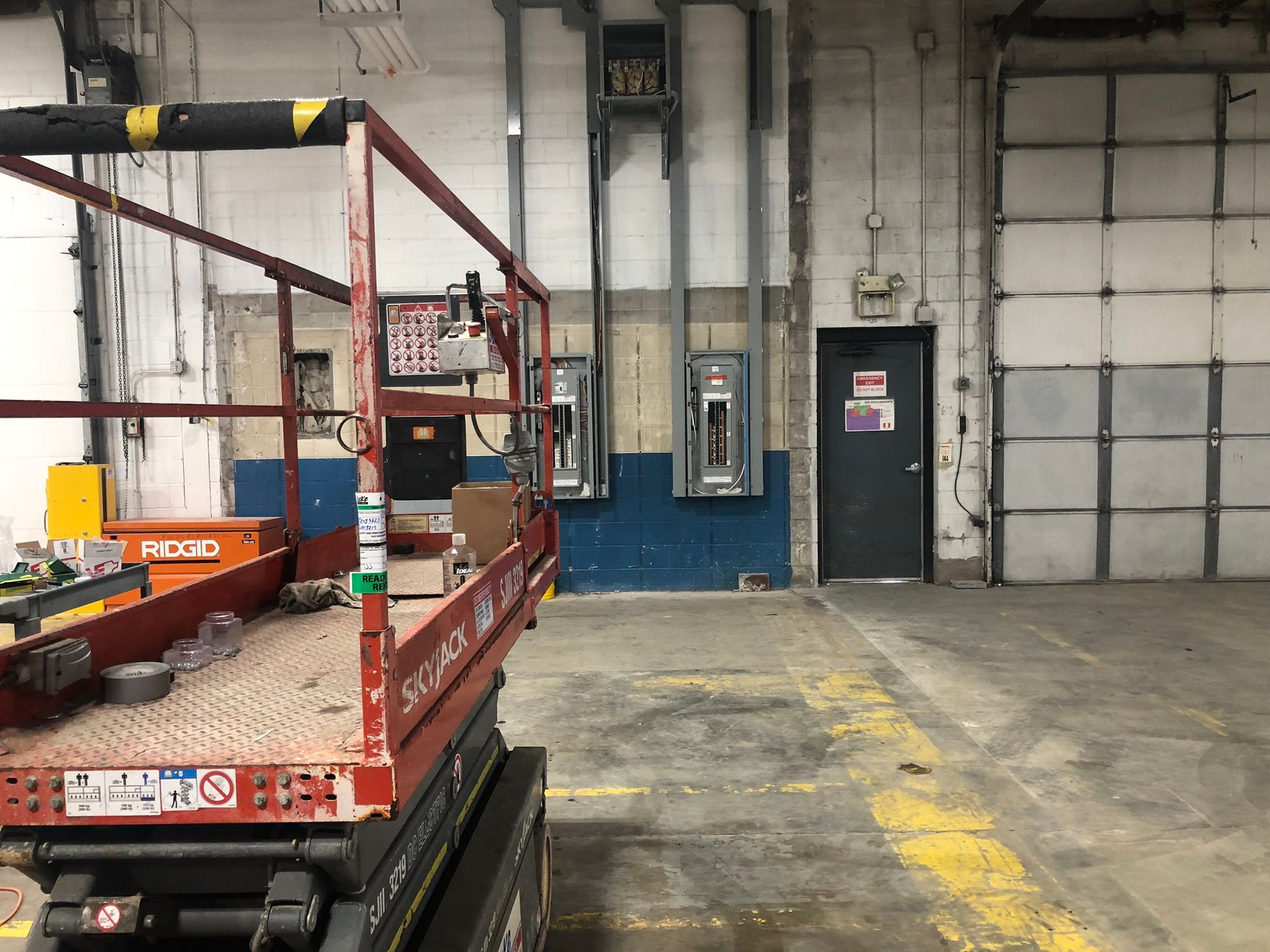 Red scissor lift in a warehouse. Electrical boxes mounted on a blue-painted wall. Door and garage door visible.