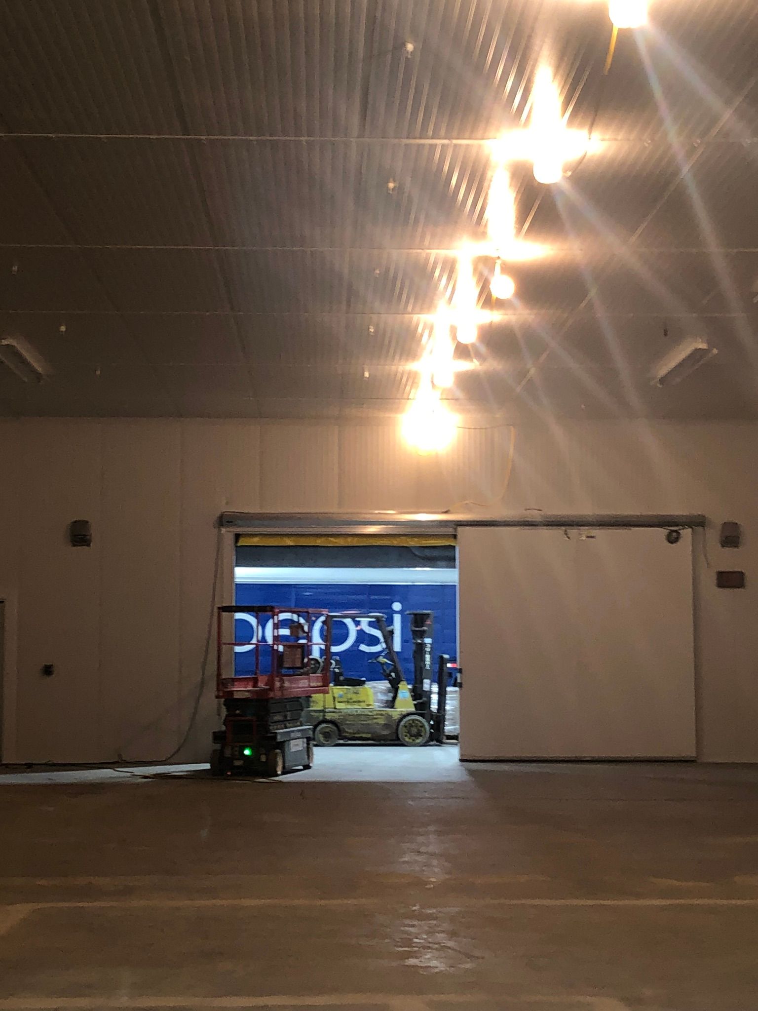 Large warehouse with open sliding door revealing Pepsi truck, forklift, and lift. Bright overhead lights.