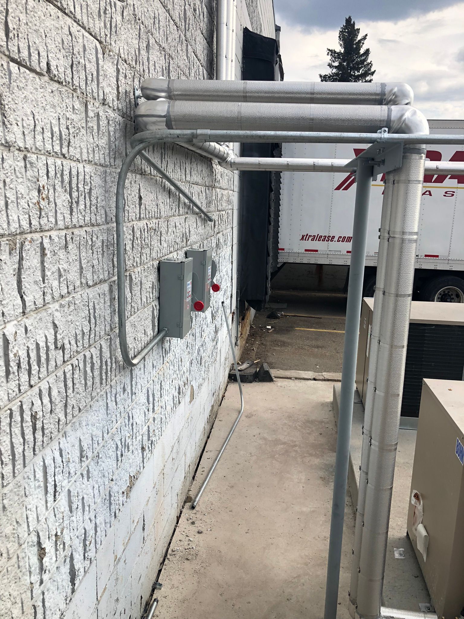 Exterior building wall with conduit and electrical boxes. Metal piping above a walkway next to a loading dock.