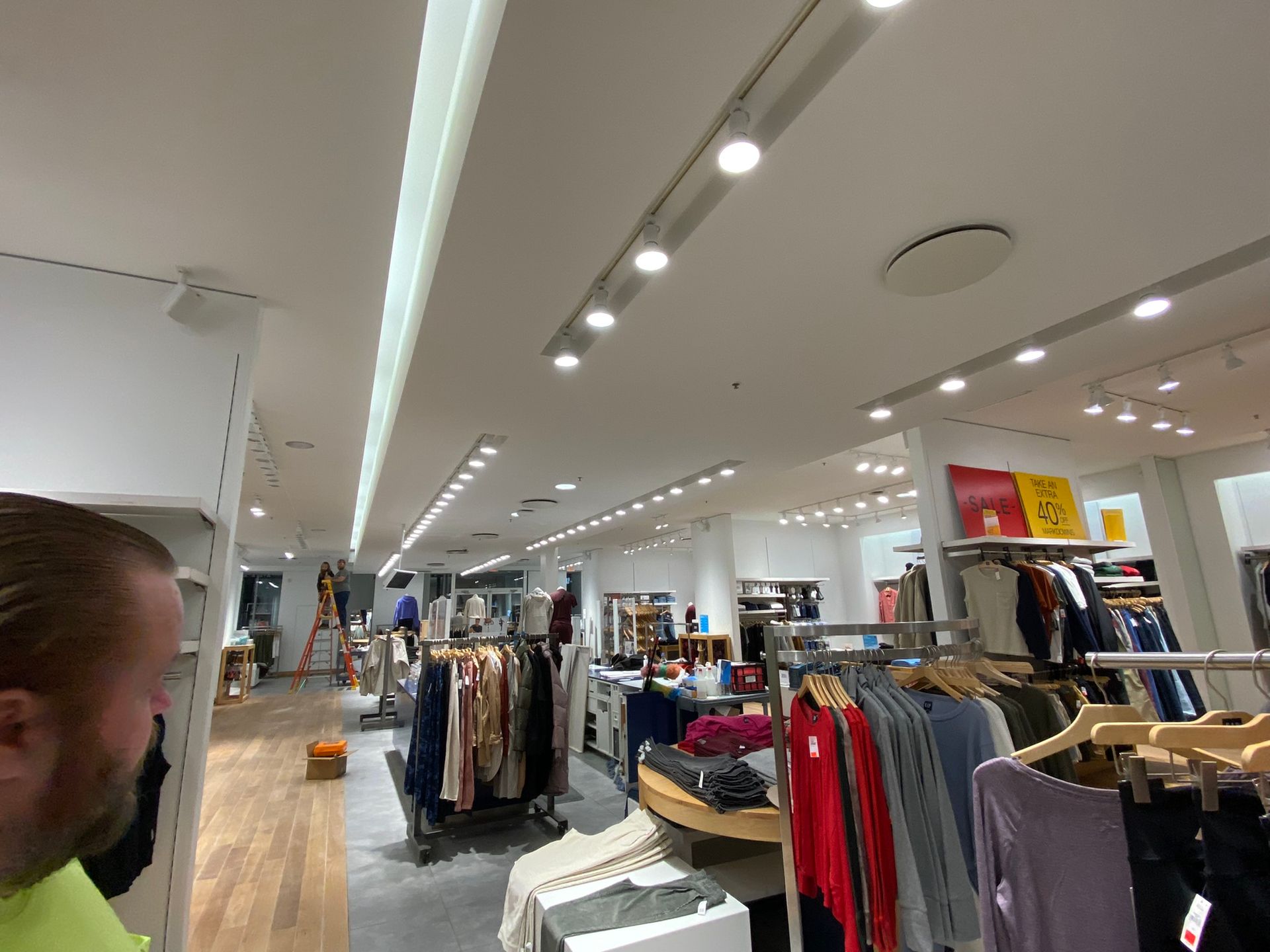 Interior view of a clothing store with racks of garments and ceiling lights. A person in a green shirt is partially visible.