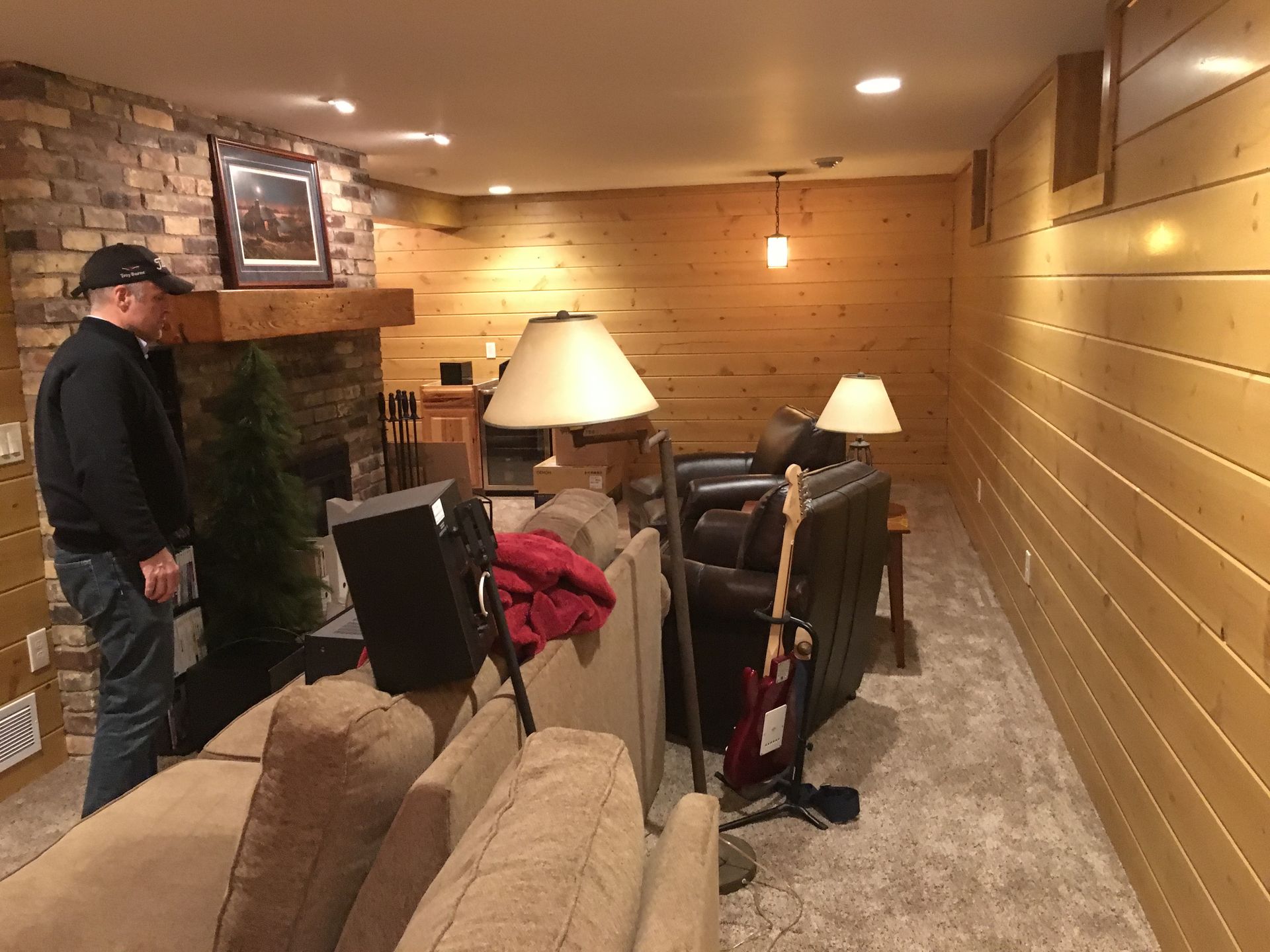 Man in basement looks at fireplace next to a sofa. Wood-paneled walls and a lamp.
