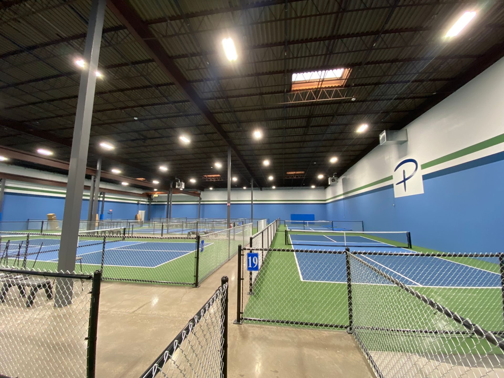 An indoor pickleball facility featuring multiple blue courts separated by chain-link fences under bright overhead lights.