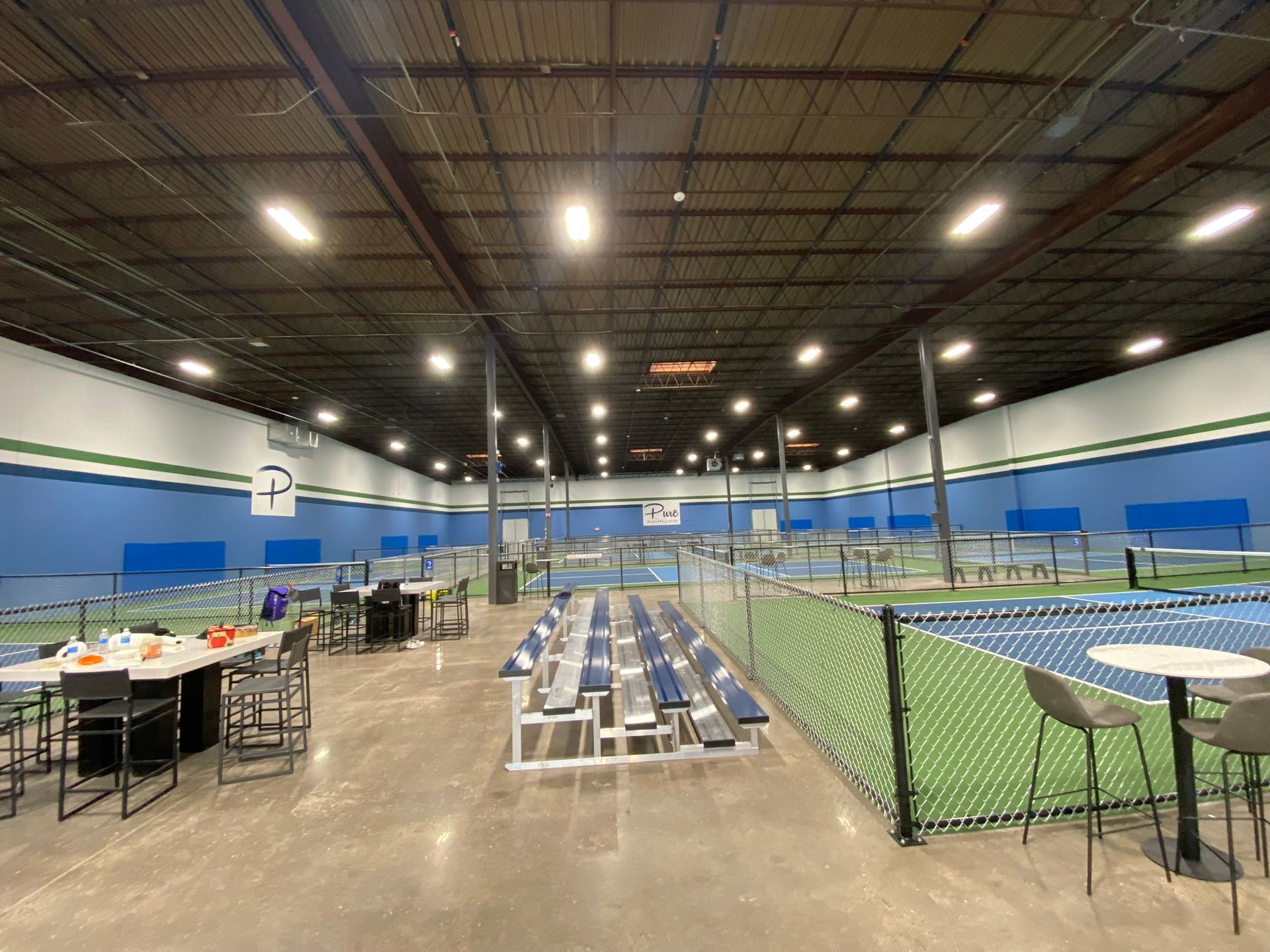 An indoor sports facility with multiple pickleball courts, spectator seating, and small tables with chairs.