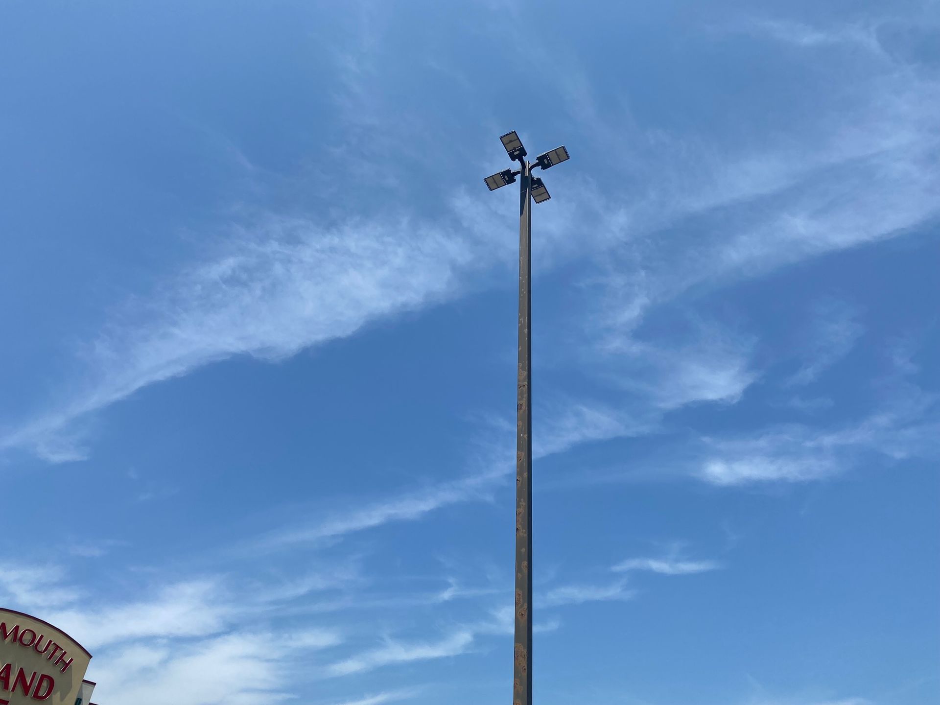 A tall metal light pole with four floodlights at the top, set against a bright blue sky with wispy clouds.